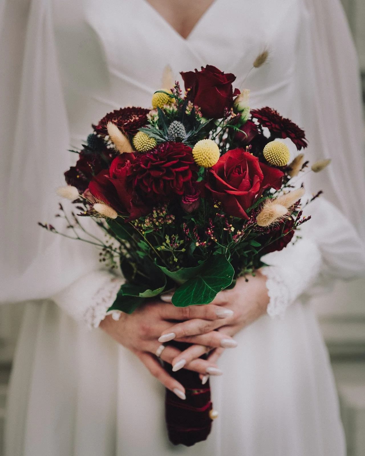 Frau in weißem Kleid hält einen roten Blumenstrauß mit Rosen, Chrysanthemen und verschiedenen Gräsern.