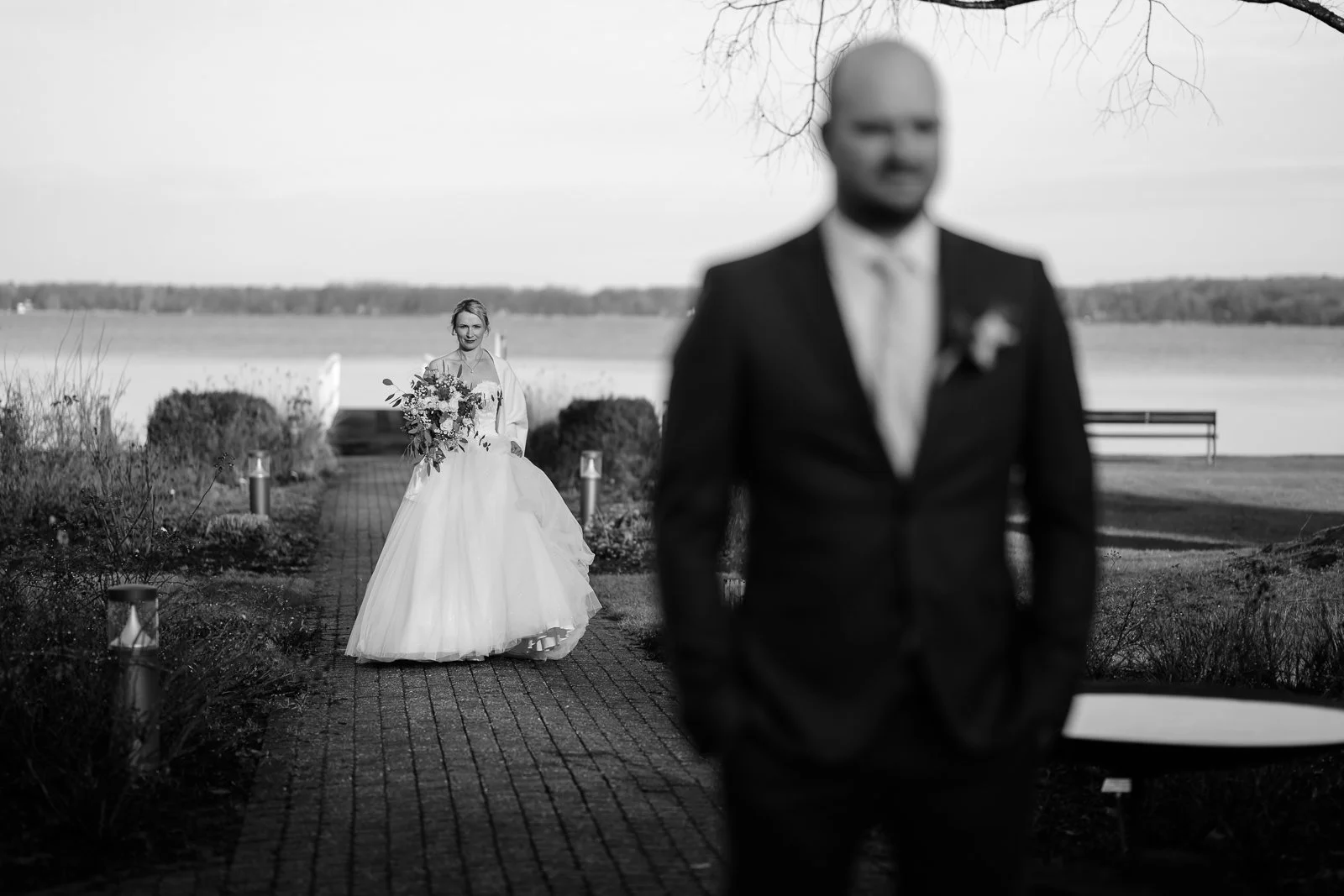 Braut in Hochzeitssaal im Hintergrund, Bräutigam im Vordergrund, unscharf, in Schwarzweiß, Hochzeit im Freien bei Wasser und Bäumen.