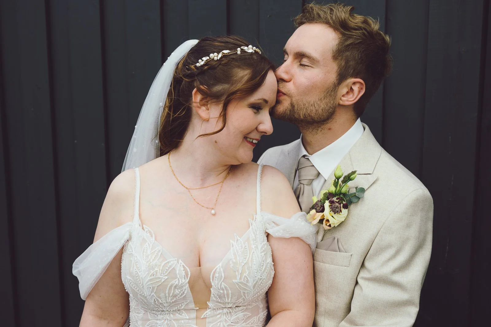 Ein Brautpaar bei einer Hochzeit, die Frau trägt ein weißes Kleid mit Spitze, der Mann trägt einen hellen Anzug mit Krawatte. Sie stehen vor einer schwarzen Holzwand, sie sind eng aneinander, sie lächeln und küssen sich leicht.