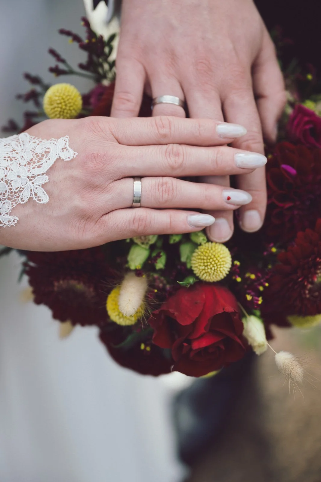 Zwei Hände mit Ringen liegen auf einem Blumenstrauß aus roten, gelben und weißen Blumen.