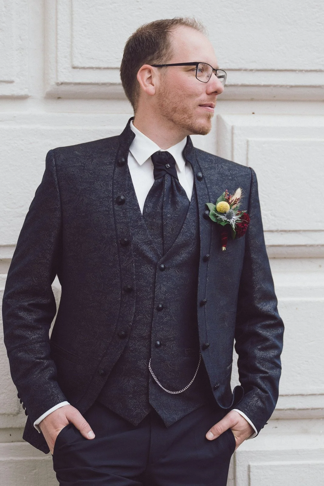 Junger Mann in formeller schwarzer Anzug mit Weste, weißen Hemd und Krawatte, trägt eine Boutonnière und steht vor einer weißen Wand.