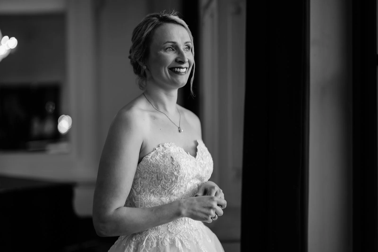 Schwarz-weiß-Fotografie einer lächelnden Frau in Brautkleid, die am Fenster steht.