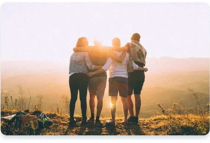 Four people standing together on a hilltop at sunset, embracing and looking at the view.