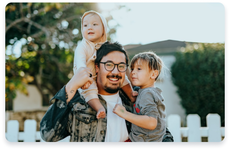 A smiling man holding two young children outdoors near a white picket fence.
