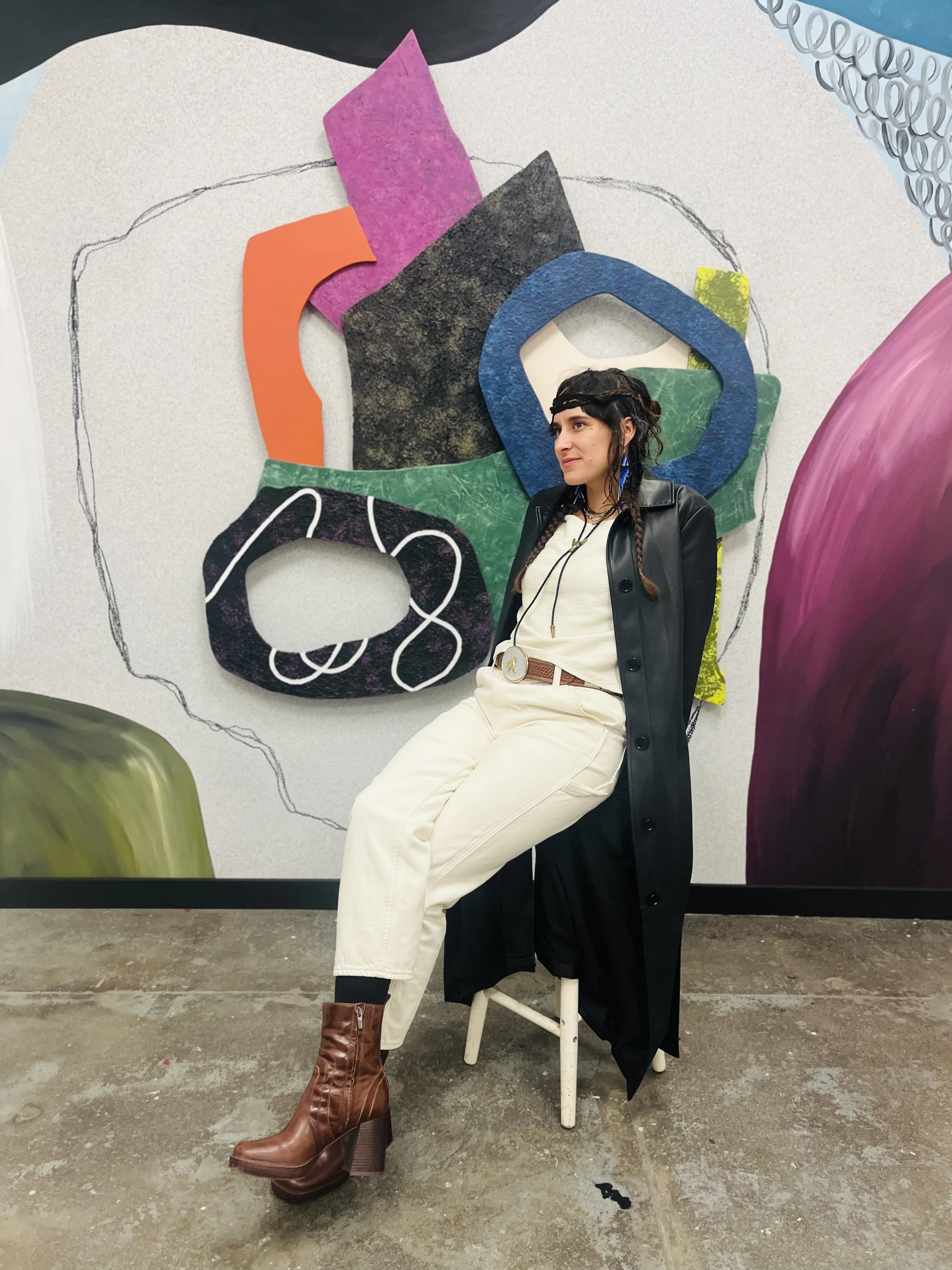 A woman with dark hair styled with braids and wearing a black jacket, a white shirt, cream-colored pants, and brown high-heeled boots, sitting on a white chair in front of an abstract art wall with colorful shapes and designs.