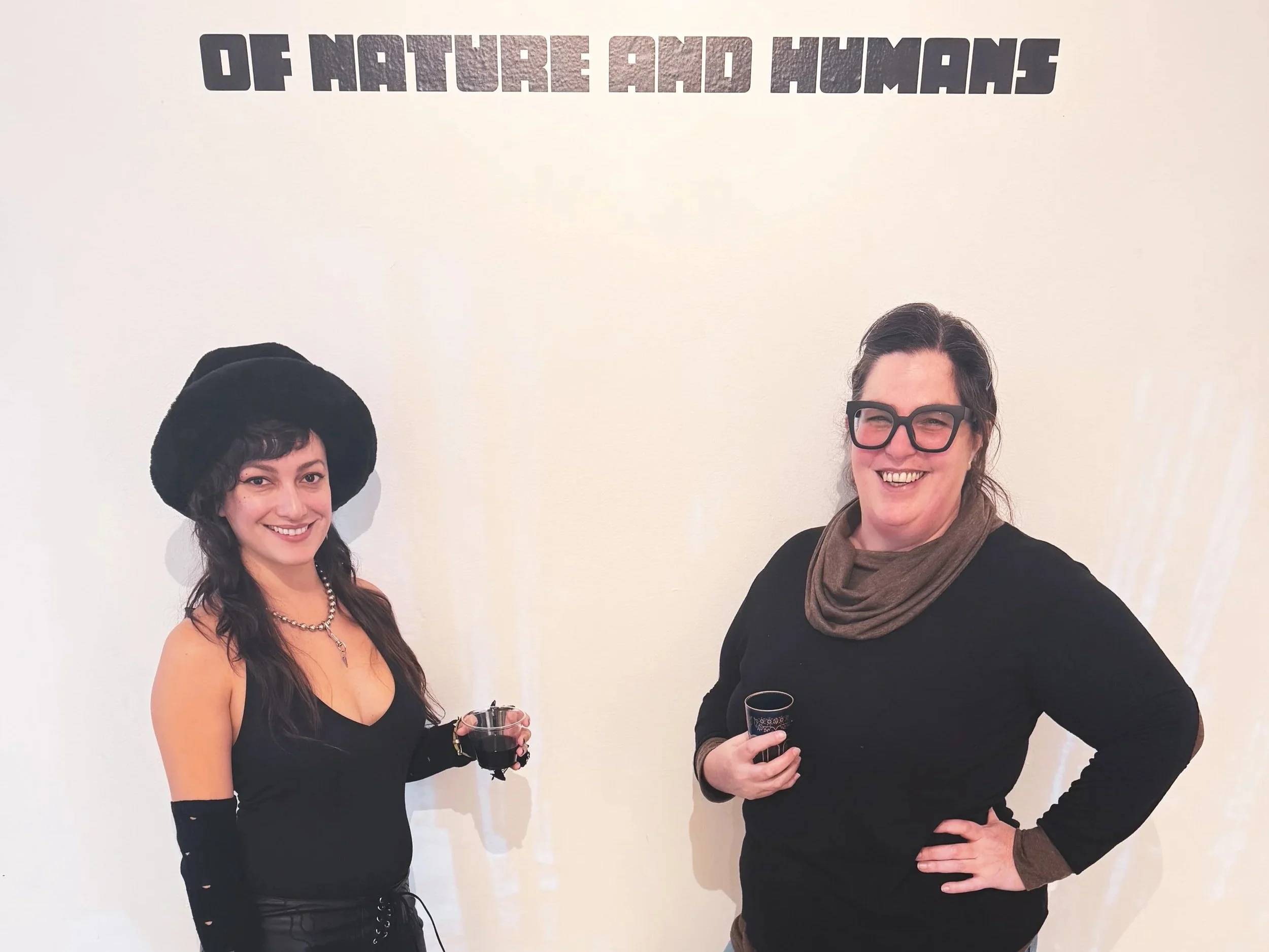 Two women standing in front of a wall with the words "OF NATURE AND HUMANS". One woman is wearing a large black hat and black dress, holding a glass of red wine. The other woman is wearing glasses, a brown scarf, and a black shirt, holding a glass of wine and smiling.