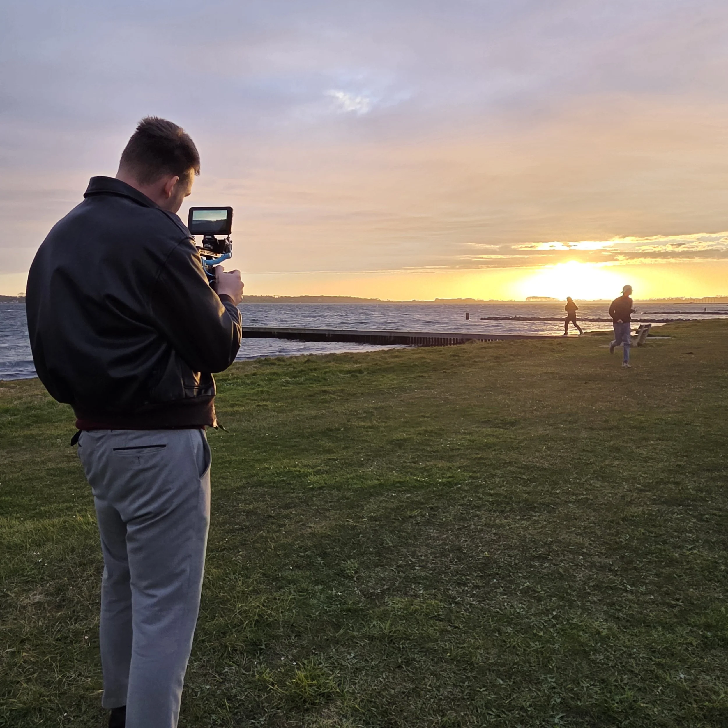 Man met een camera die de zonsondergang boven water fotografeert, omringd door andere mensen op het gras bij de rivier.