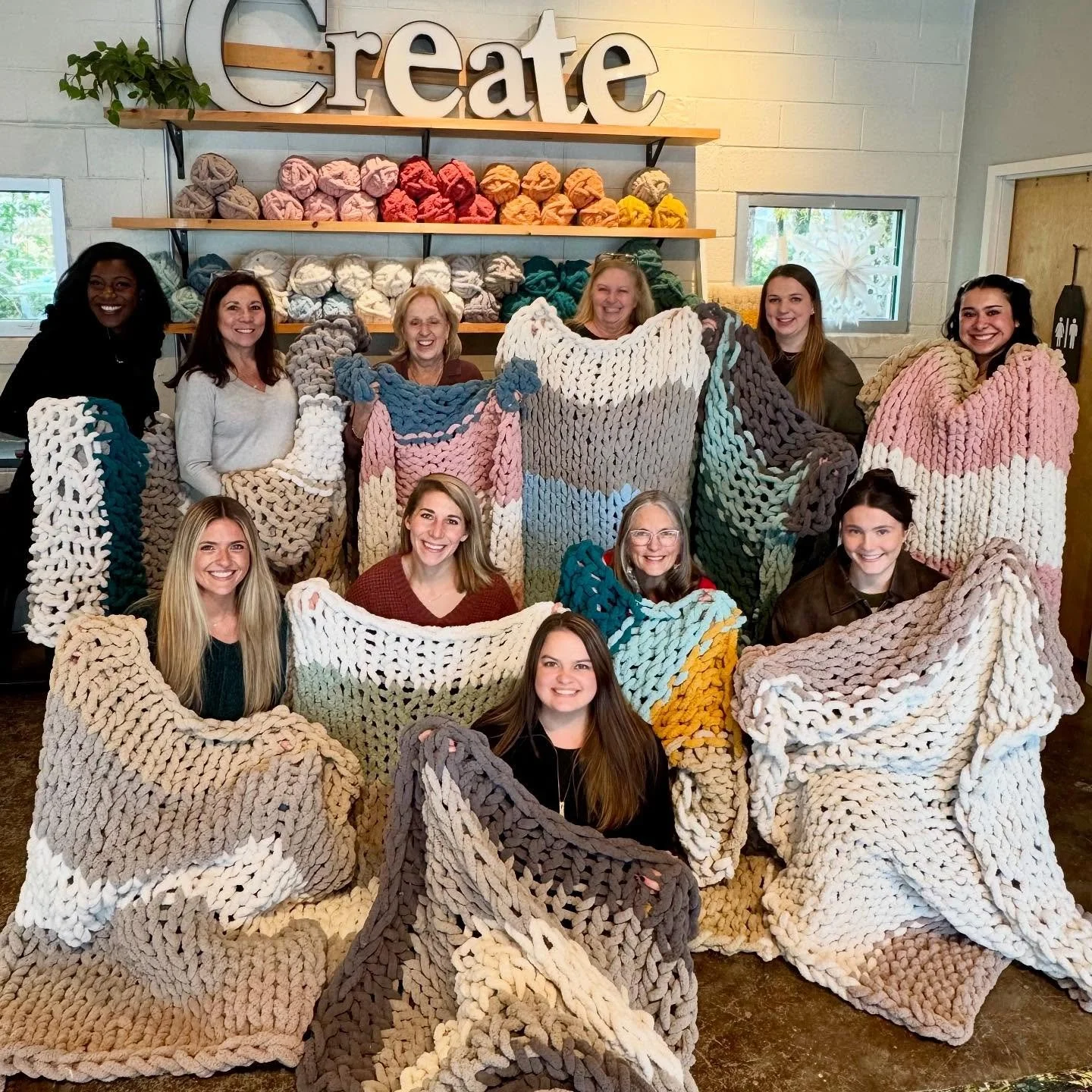 Eight women standing indoors, wearing large, crocheted blankets of various colors, smiling. Behind them, a large sign spells 'Create' on a wall.
