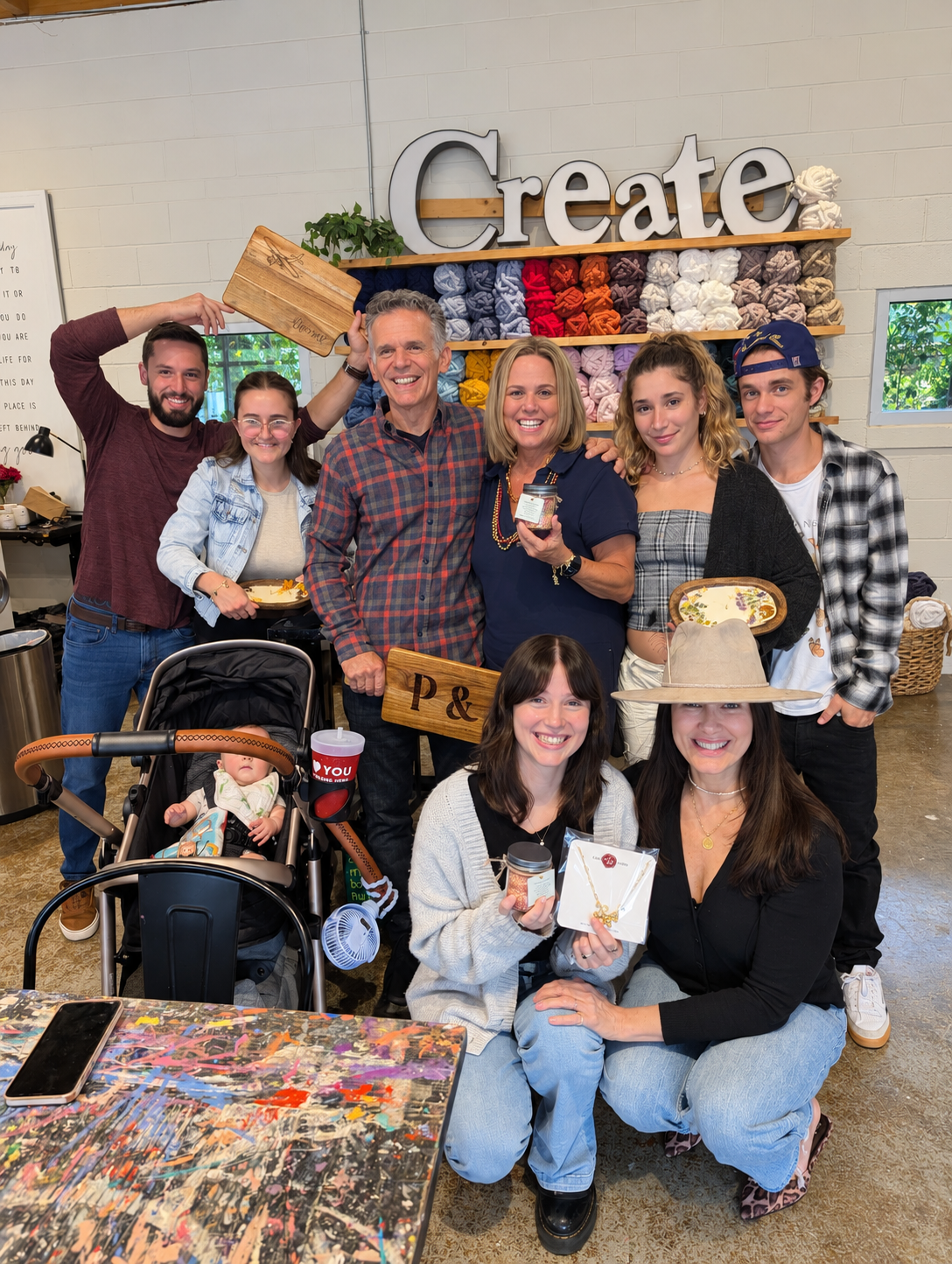 Group of women at a crafting table in a craft store, holding jewelry and smiling, with craft supplies and completed projects on shelves in the background.