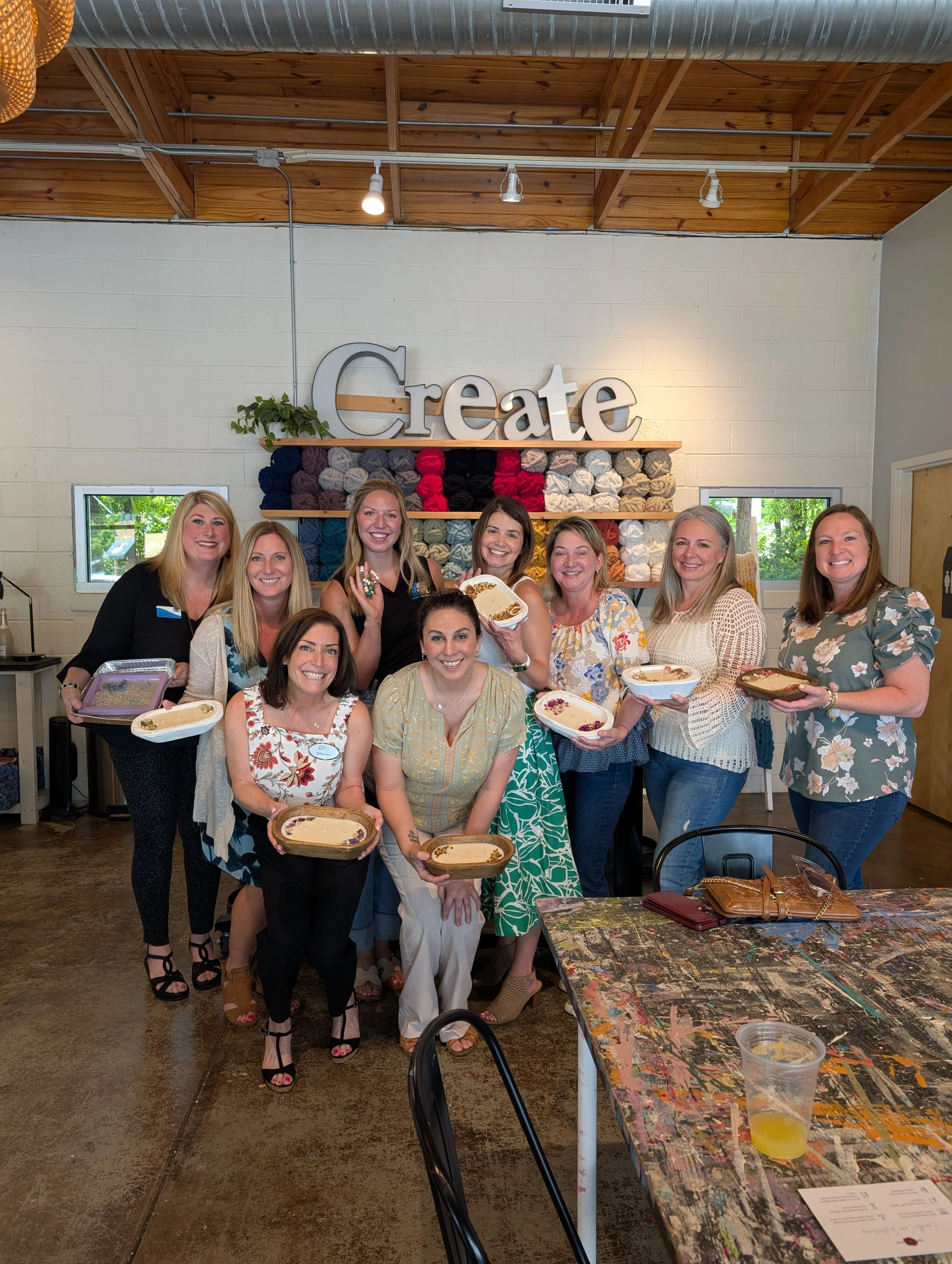 Group of women at a craft or pottery class holding painted ceramic dishes in front of a wall with a large 'Create' sign.