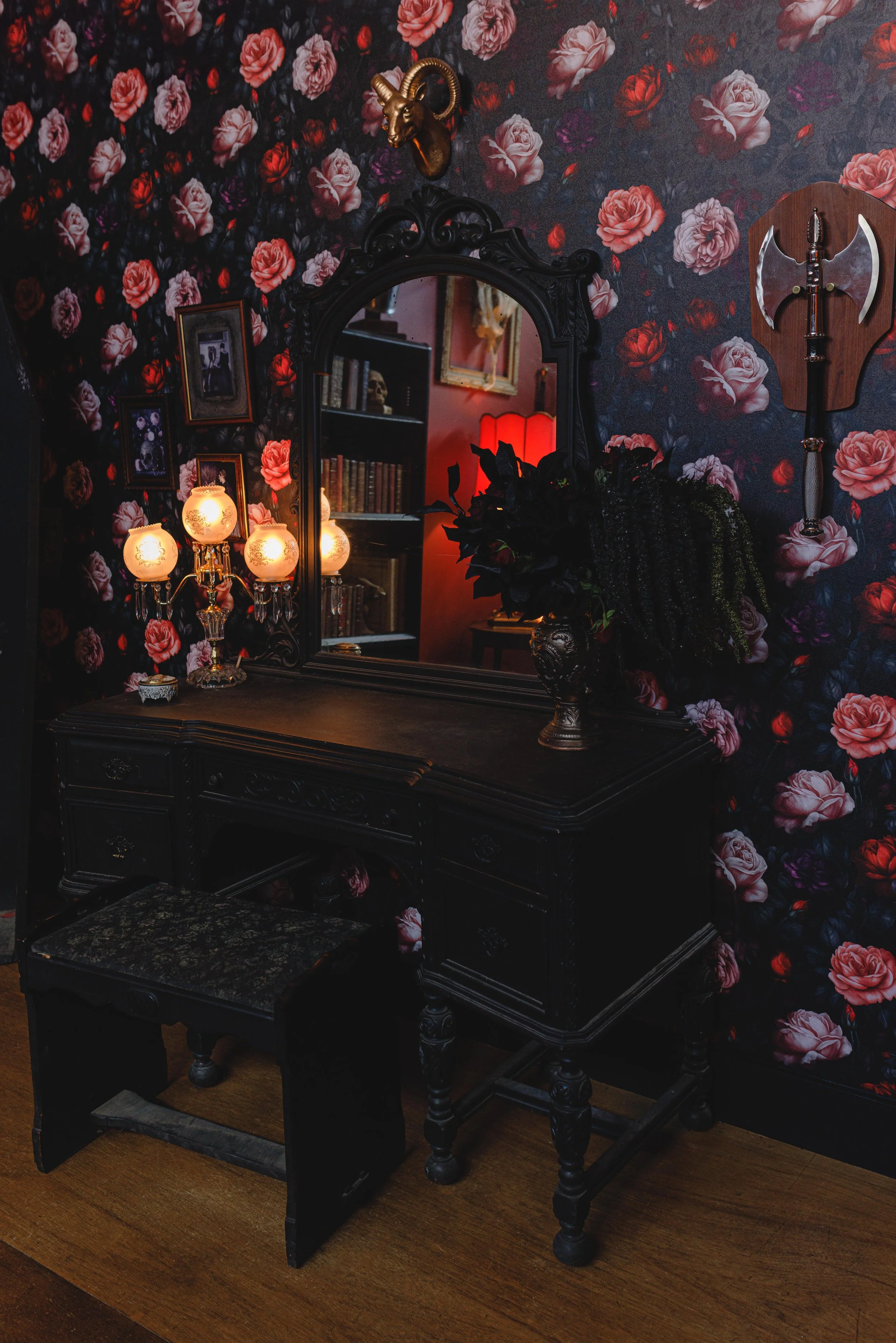A black vintage vanity table with a mirror, decorated with a vase of dark flowers, a lamp with four globe-shaped lights, and framed photos, on a wooden floor with floral wallpaper background.