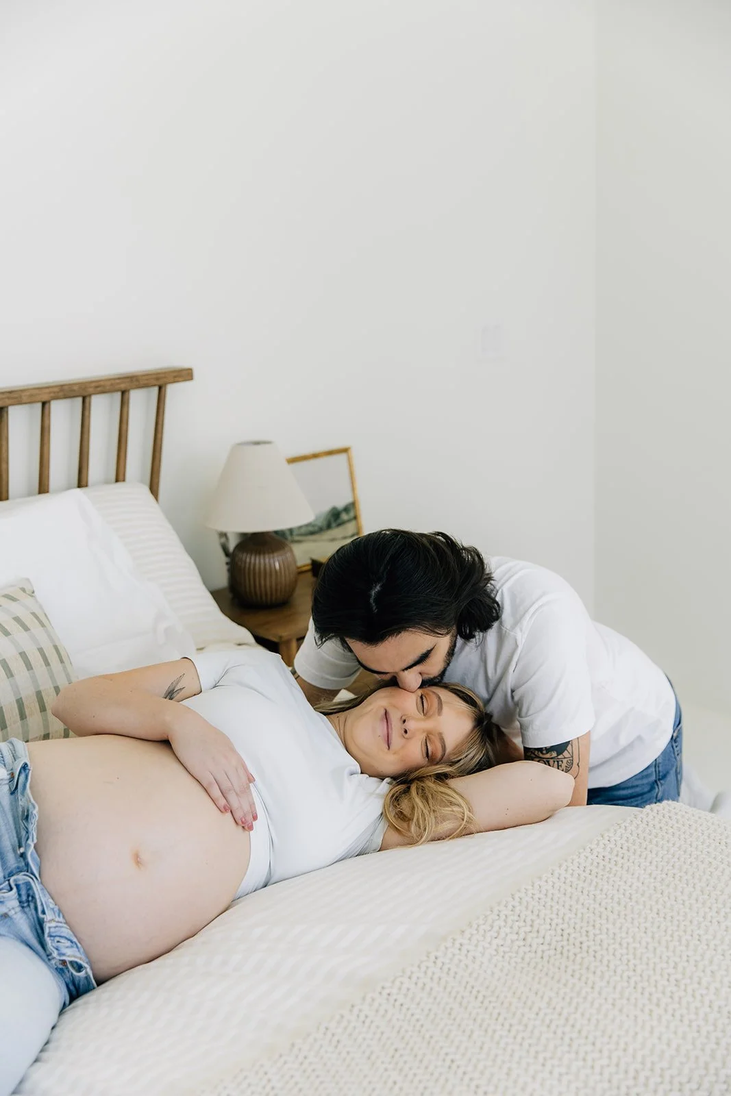 A man kissing a pregnant woman on the forehead while lying on a bed in a cozy bedroom.