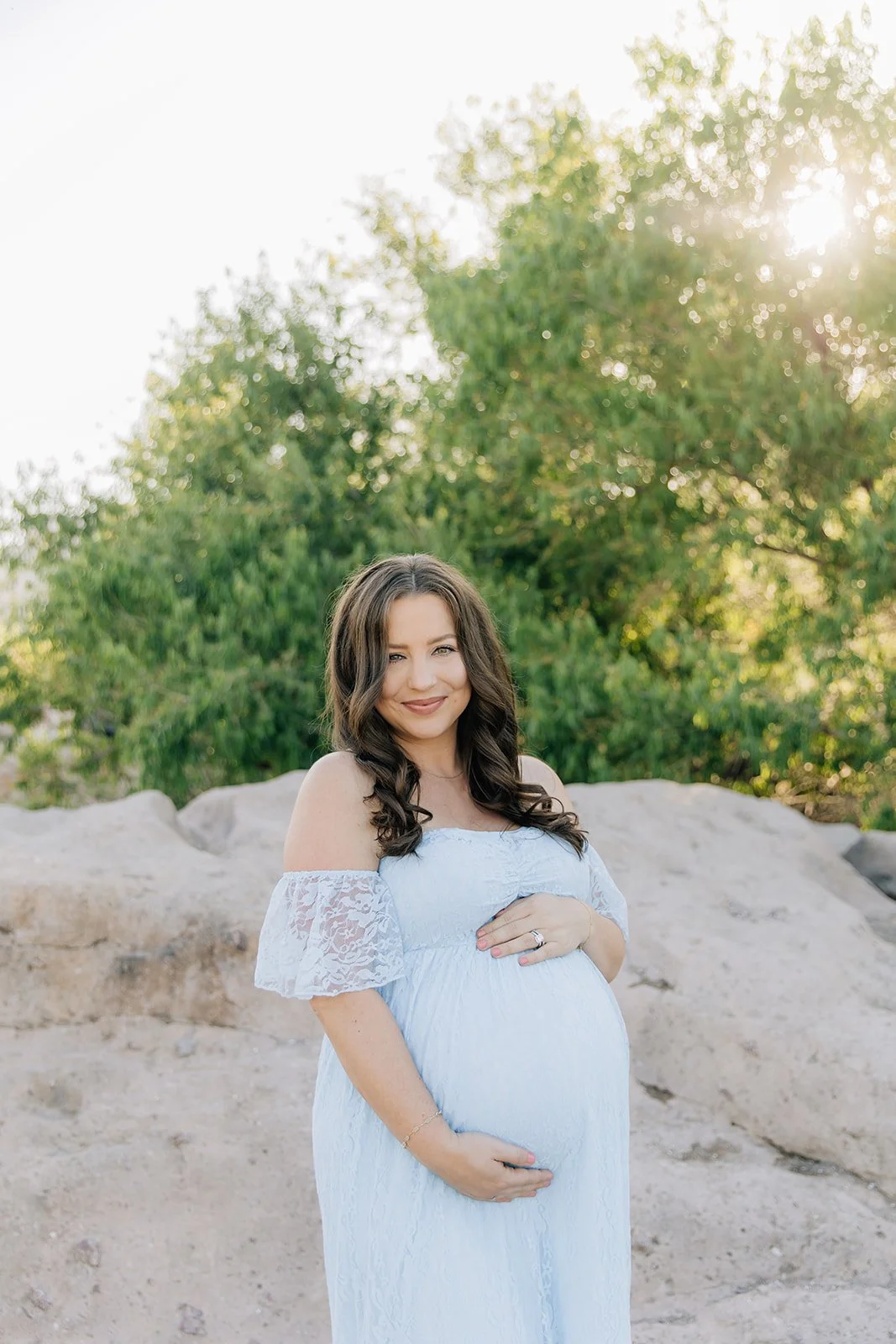 Pregnant woman in a white lace dress standing outdoors with trees in the background during sunset, smiling and cradling her belly.