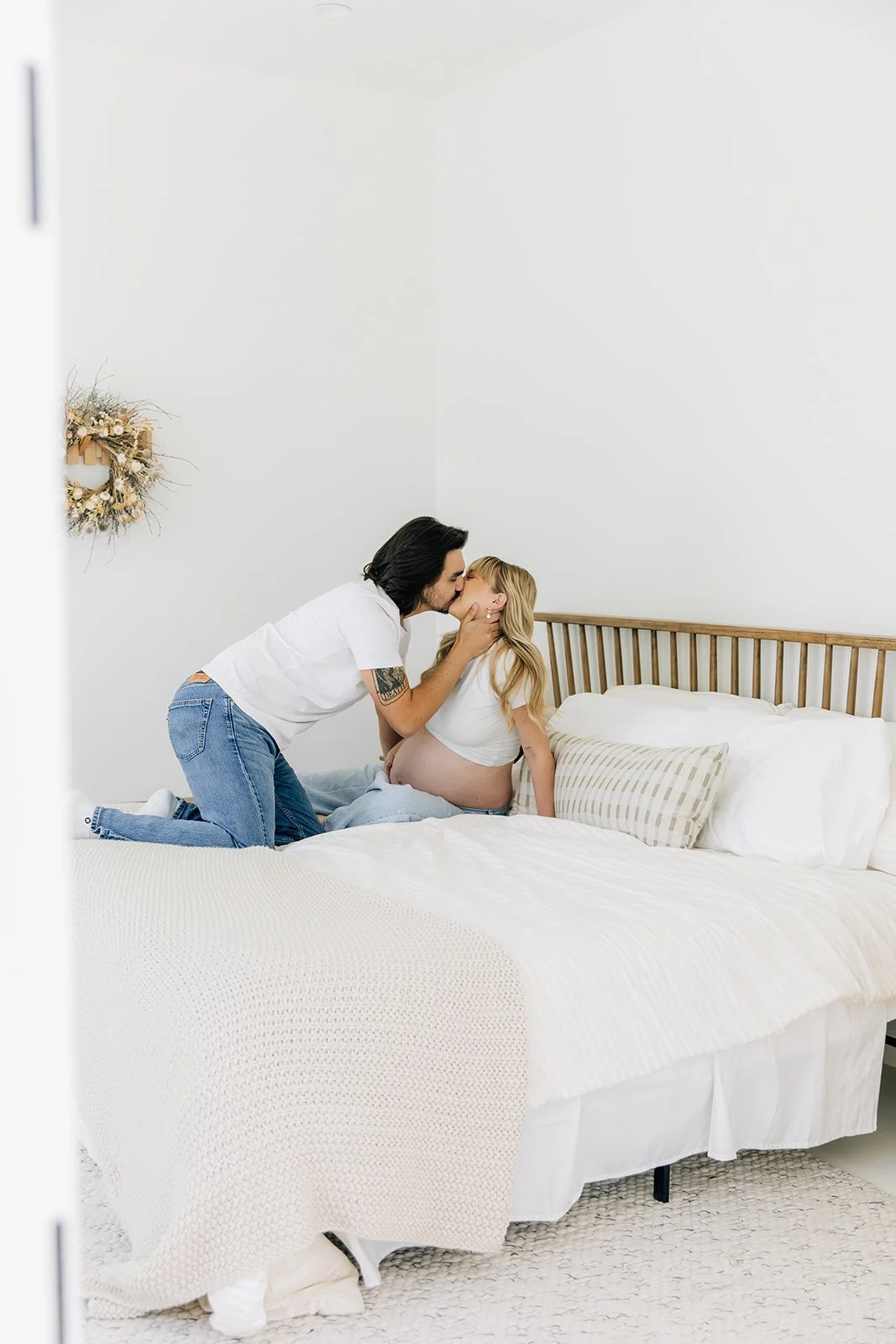 A couple sharing a kiss in a bedroom during a maternity photoshoot, with the pregnant woman sitting on a bed.