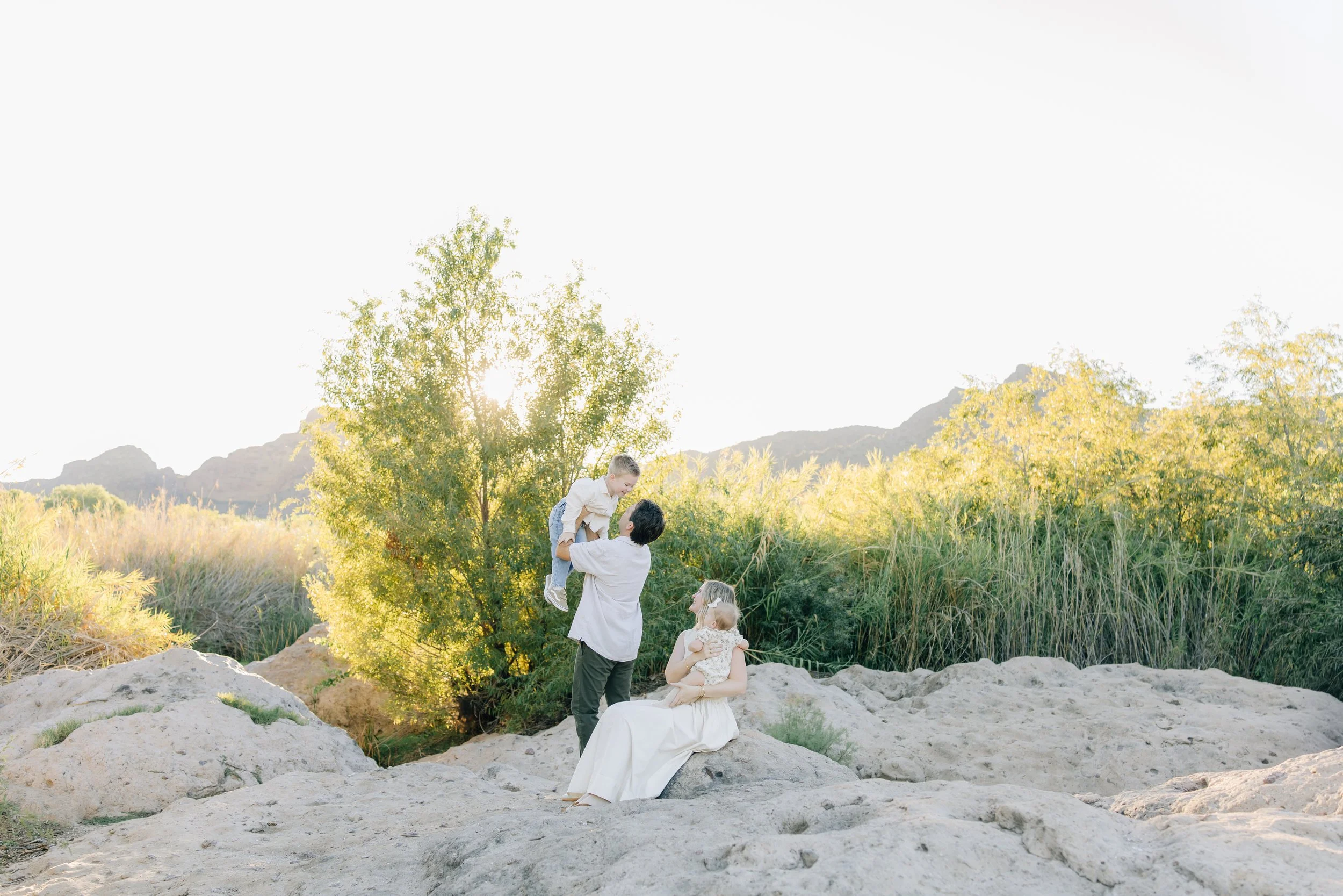 A happy family of four in the Arizona desert, with a man standing and woman sitting holding a baby on a rocky area and a young boy being lifted in the air. Tall plants and Arizona desert shrubs are in the background.
