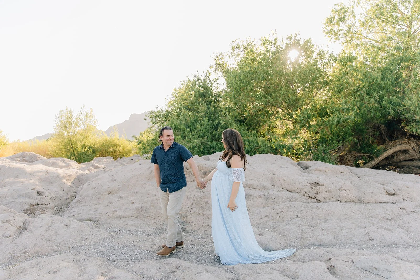 A man and a pregnant woman holding hands on rocky terrain with greenery and sunlight in the background.