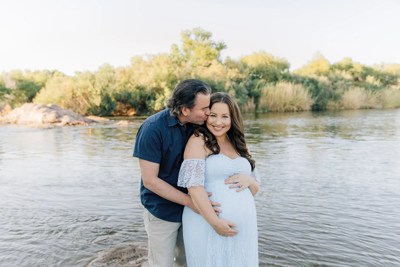 A man and woman standing in a river, with the man kissing the woman's cheek. The woman is pregnant and smiling at the camera. There are trees and bushes in the background, and the sky is clear.