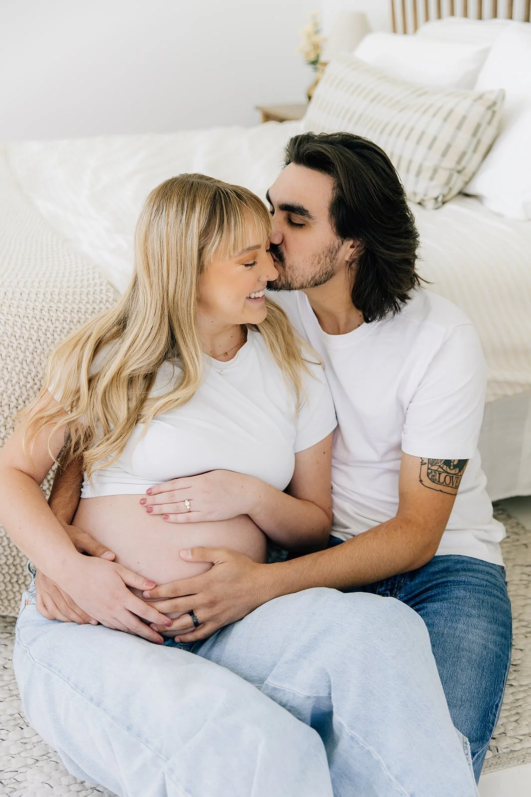 A pregnant woman and a man are sitting on the floor, with the man kissing the woman's forehead. The woman is smiling and resting her hands on her belly, while the man is holding her belly with both hands. They are in a cozy bedroom.