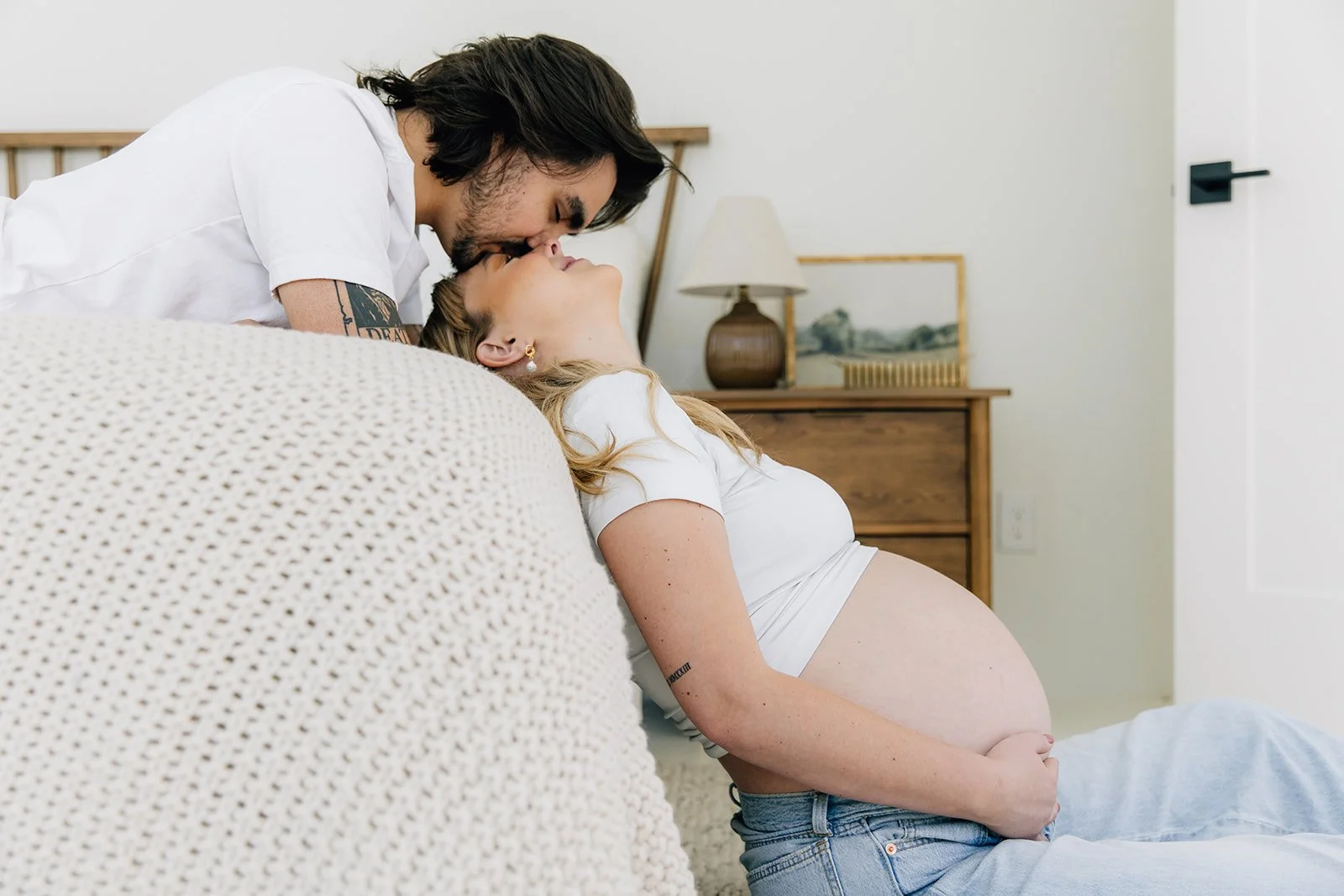 A couple sharing a kiss in a bedroom; the woman, pregnant, is sitting on the floor, leaning back against a beige couch, with her hand on her belly, while the man leans over her, holding her head.