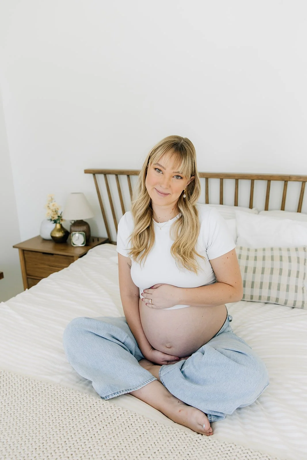 Pregnant woman sitting on a bed in a bedroom, smiling and looking at the camera.