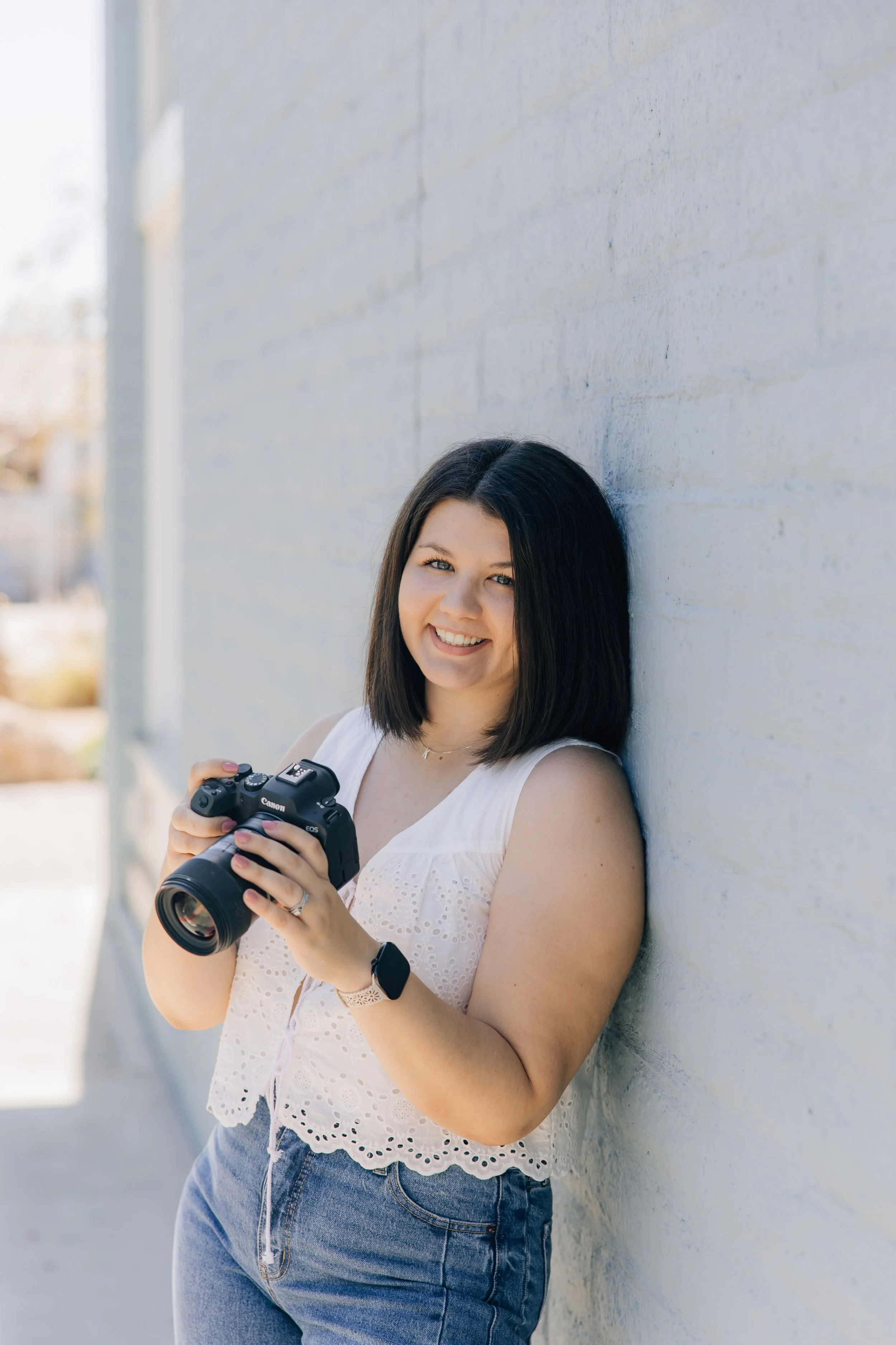 Women leaning against the wall while holding a camera up in her hands smiling at the photographer.