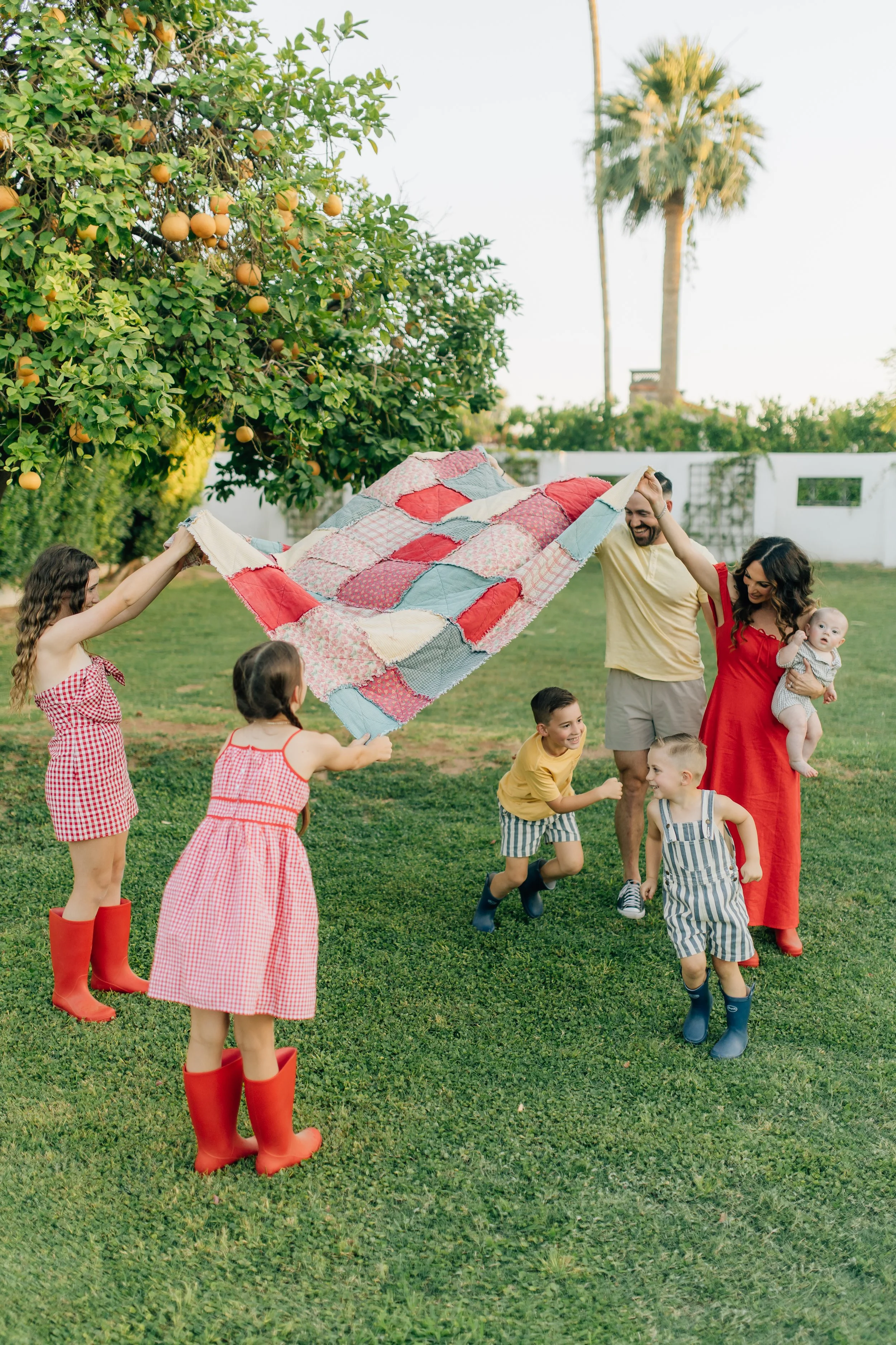Family of seven playing in their yard. Parents and two older daughters lifting up a blanket while two brothers run under.