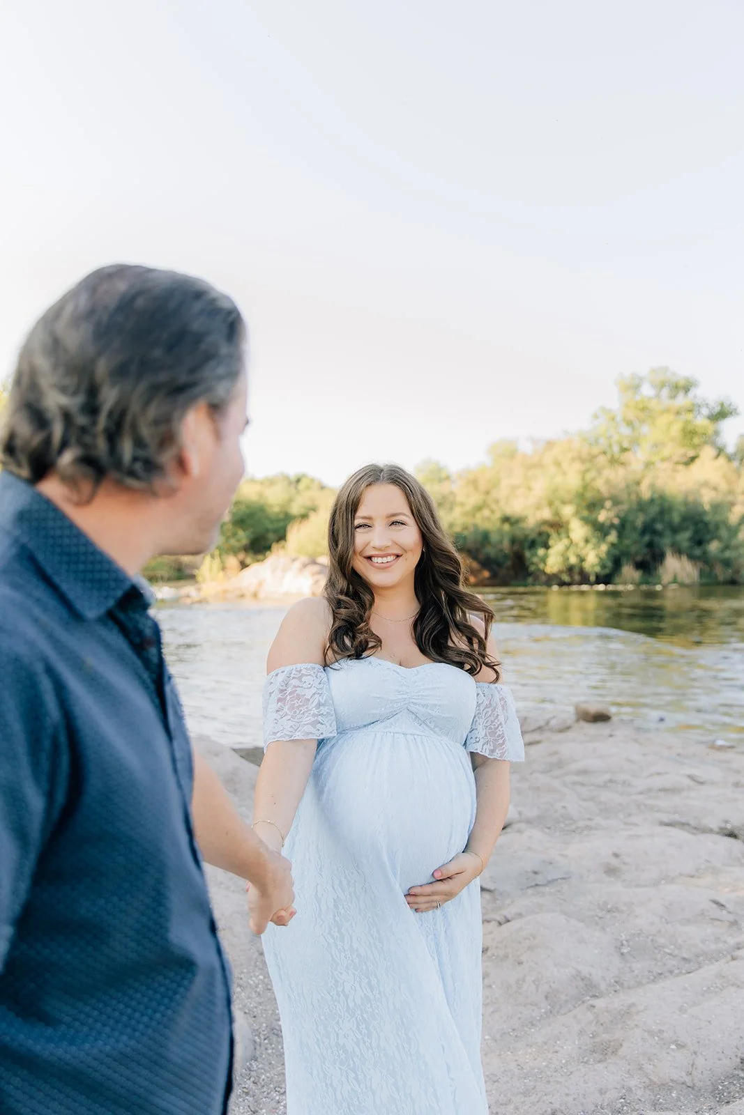A pregnant woman in a white dress standing by a river, smiling at a man holding her hand during sunset.