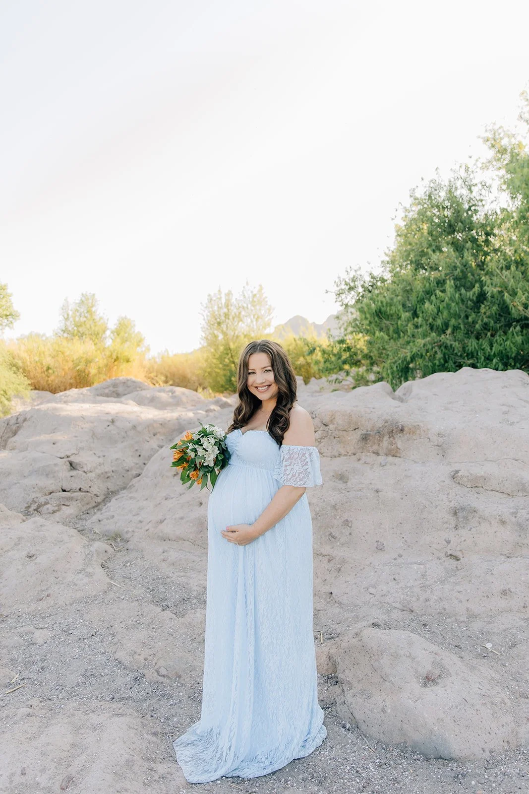 A pregnant woman in a white dress holding a bouquet of flowers, standing outdoors on a rocky landscape with green trees in the background, smiling at the camera.