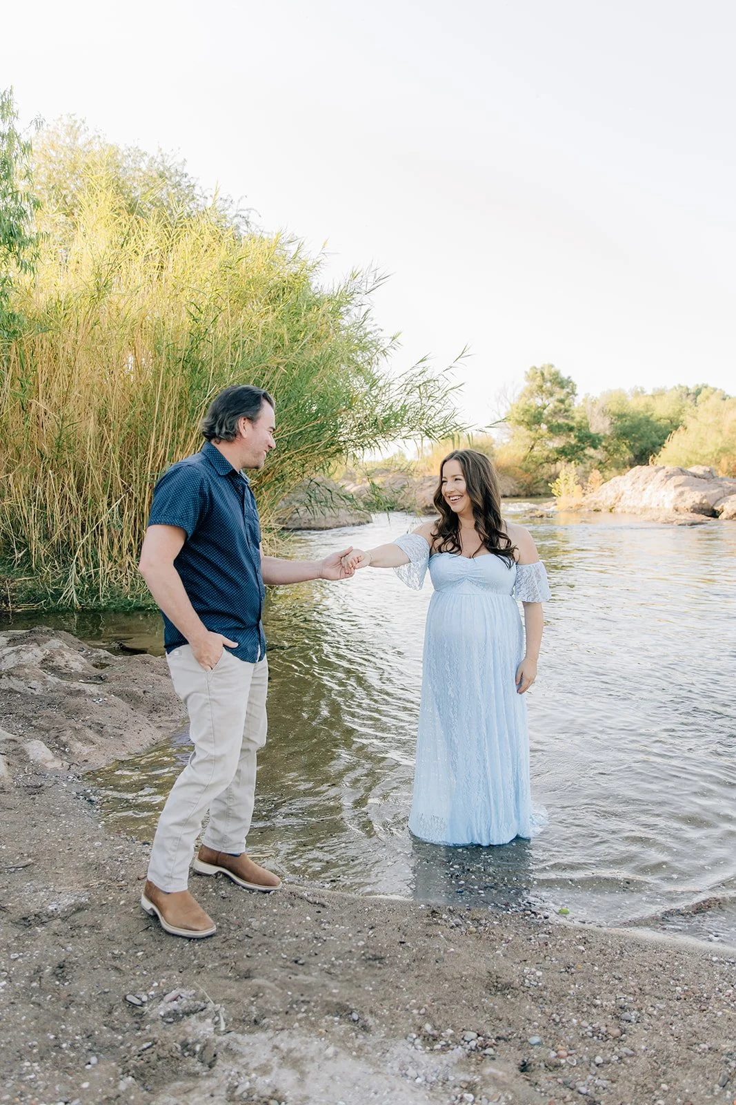 A couple standing by a river, with the woman touching the man's hand, both smiling. The woman wears a white dress, and the man wears a blue shirt and beige pants.