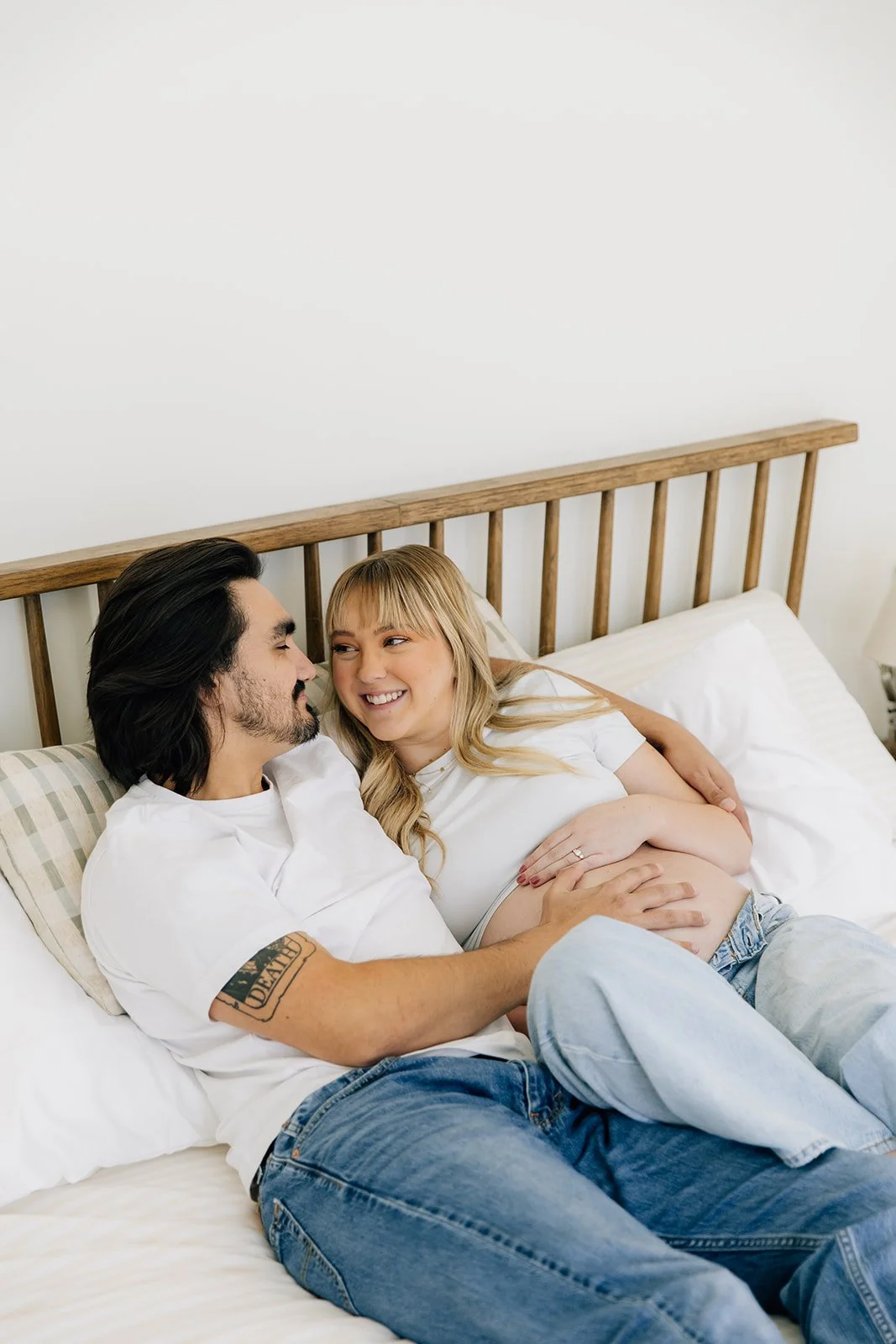 A happy couple sitting on a bed, with the woman showing her pregnant belly, smiling at each other