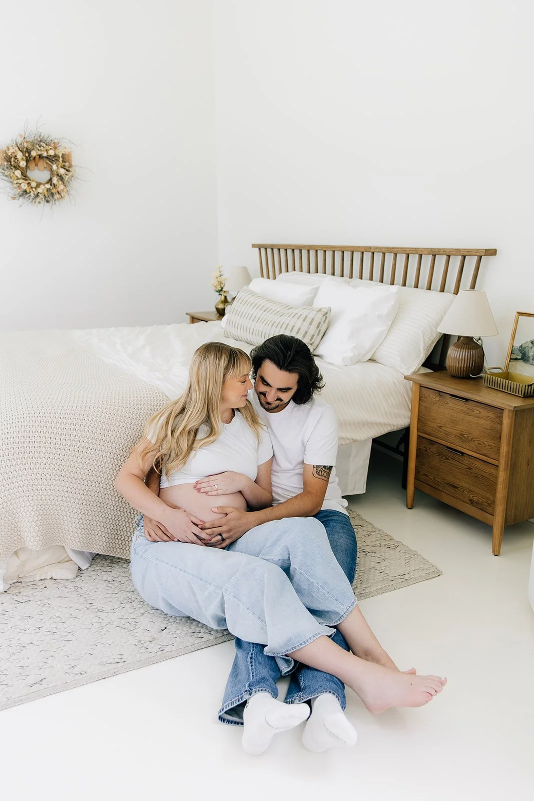 A couple sitting on the floor in front of a bed, with the pregnant woman resting her belly on the man's lap, in a bright bedroom with white walls, wooden furniture, and minimal decor.