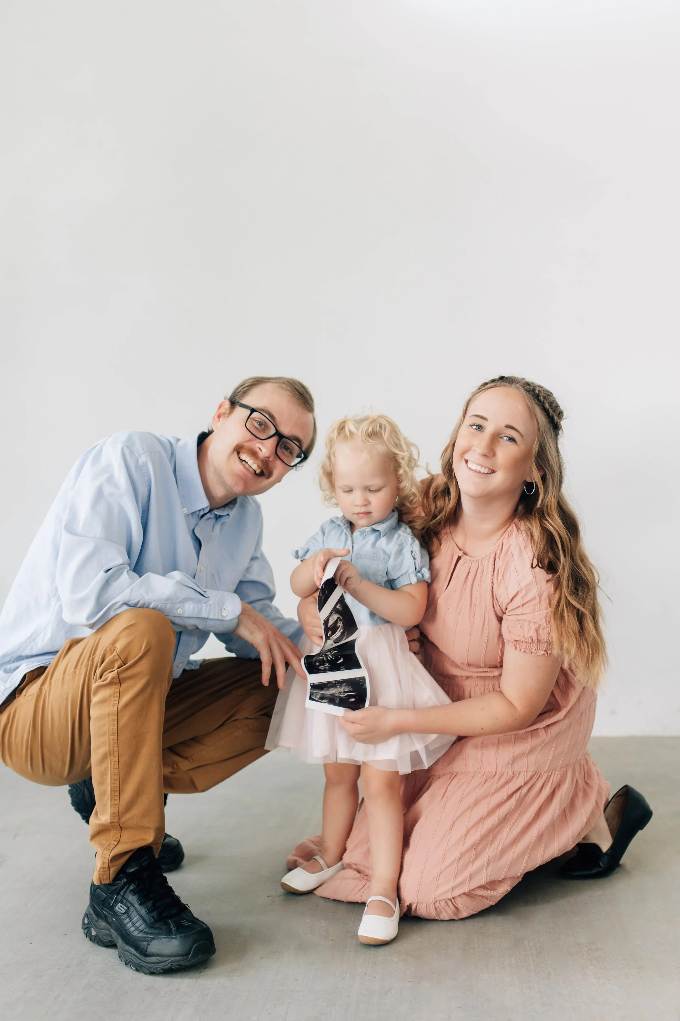 A family of three: a father, mother, and young girl, all smiling and holding ultrasound images, in a bright, minimalistic room.