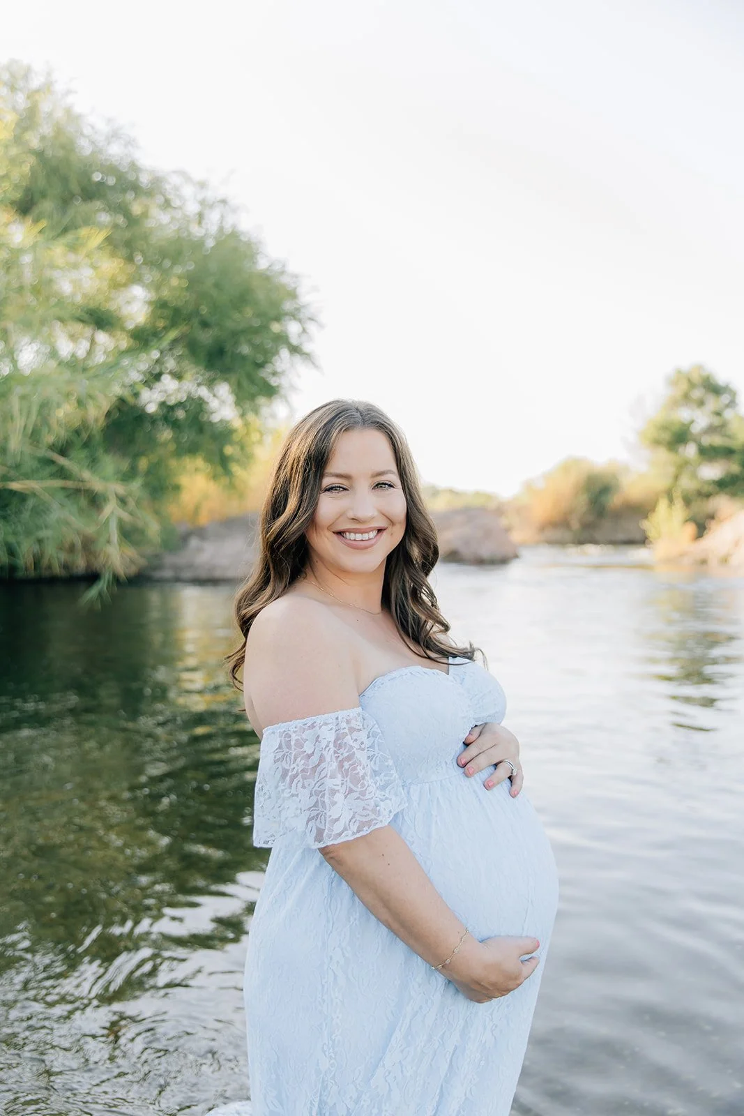 Smiling pregnant woman in a white lace off-shoulder dress standing by a river with trees and rocks in the background during daylight.