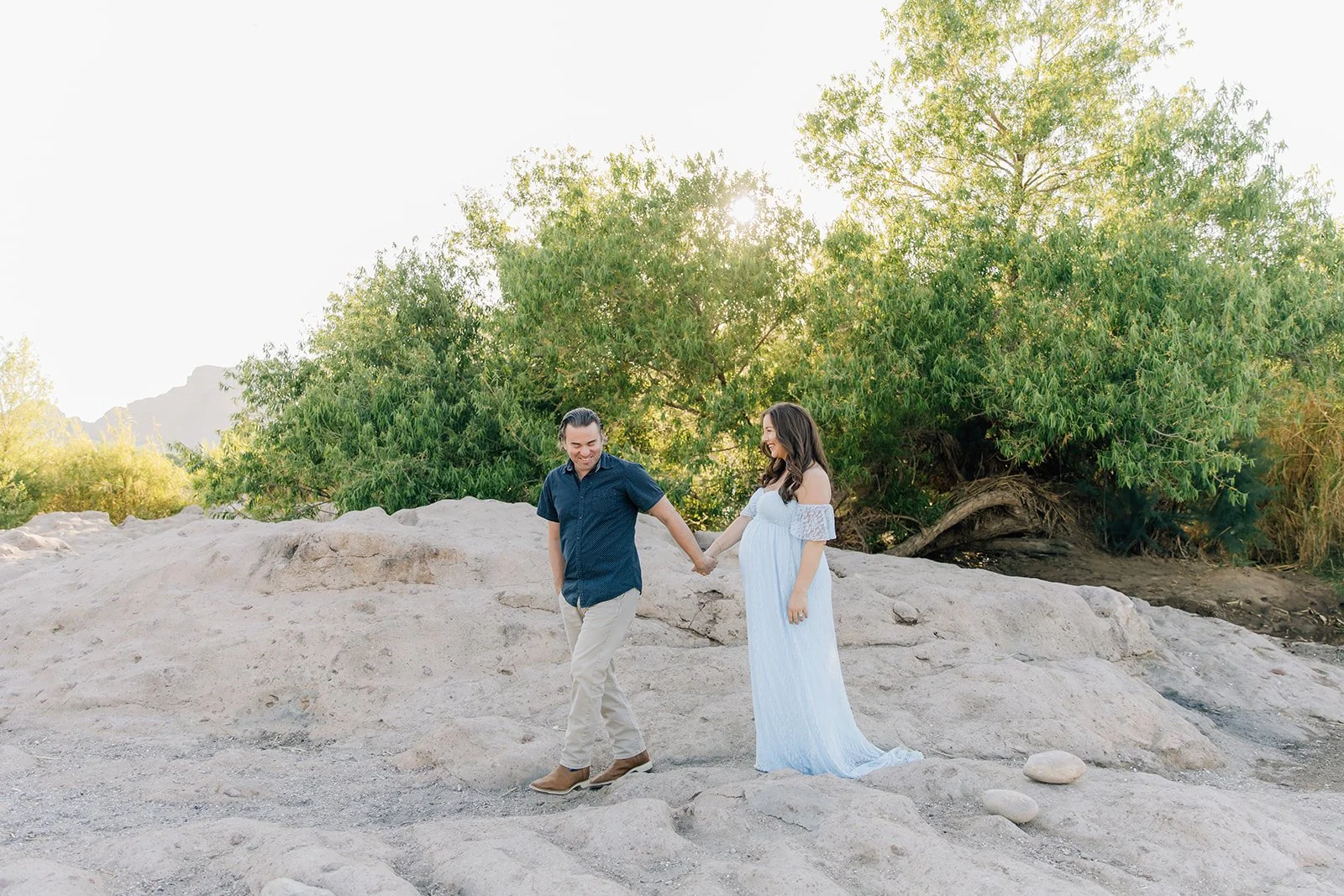 A pregnant woman in a white dress holding hands with a man in a dark shirt and beige pants on rocky terrain with green trees and the sun shining through the leaves