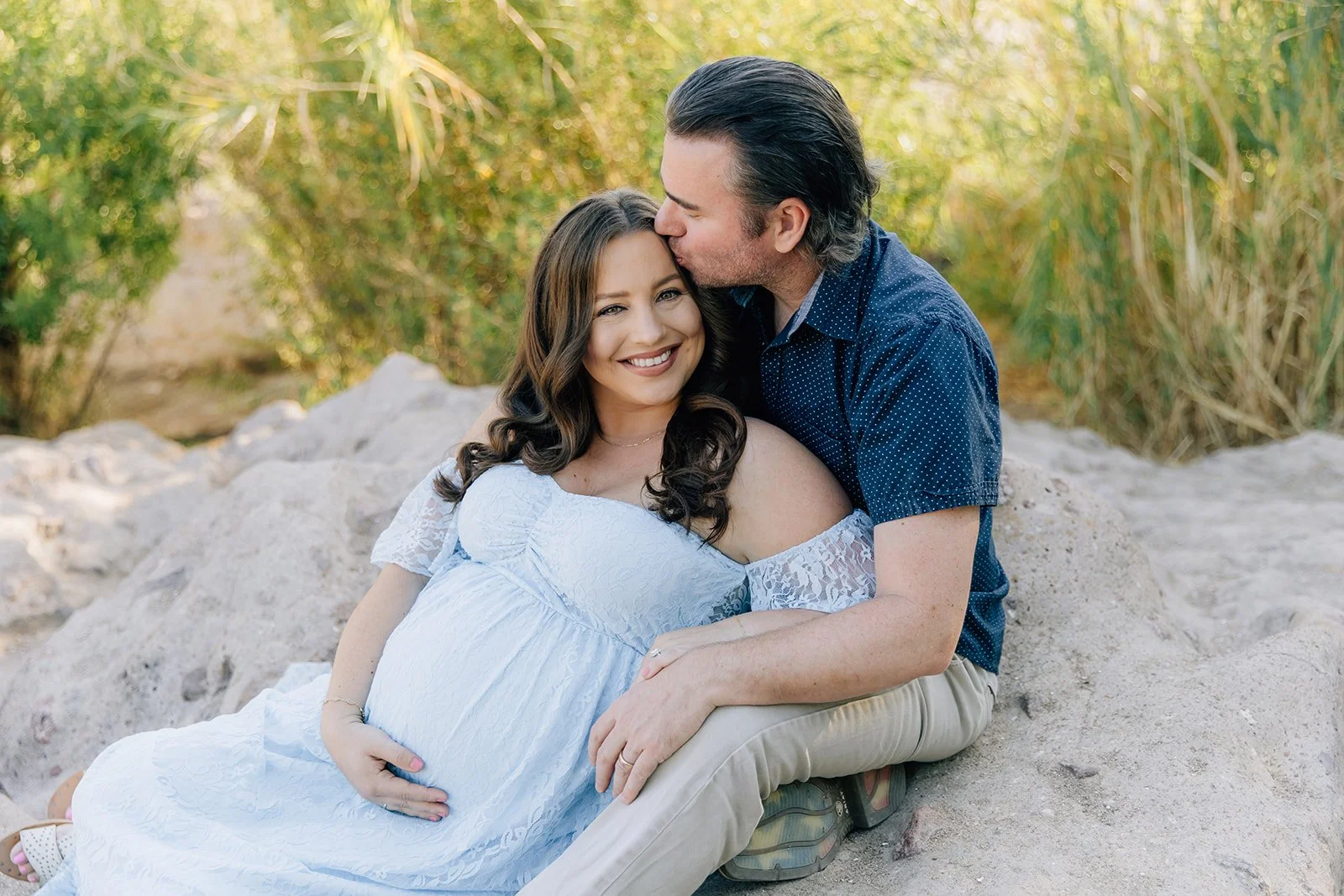A couple sitting on rocks in nature, the woman is pregnant, and the man kisses her forehead while she smiles at the camera.