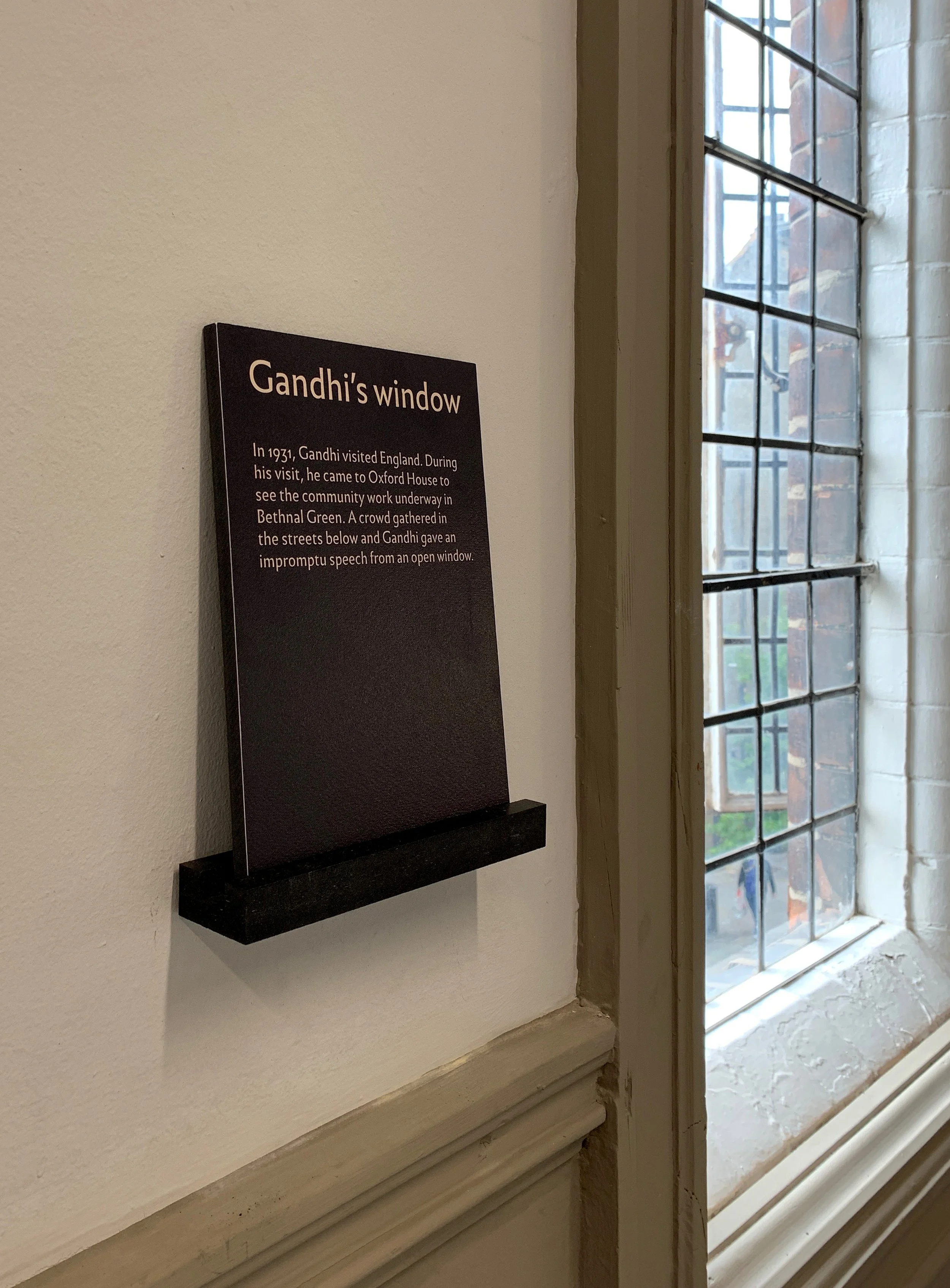 A black exhibition label with white text sits on a small black shelf next to a paned window. It describes Mahatma Gandhi giving a speech from this window in 1931.