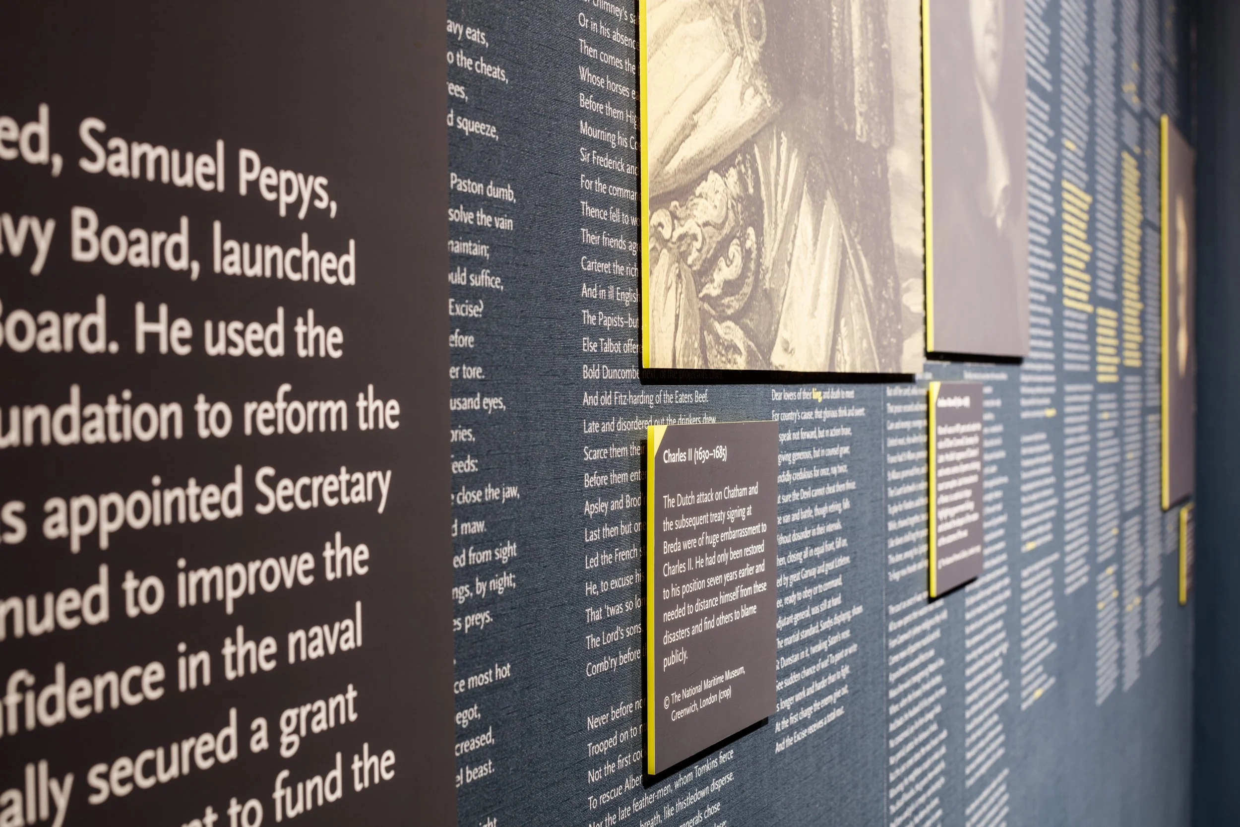 Close-up of text on a blue exhibition wall, featuring white printed text about Samuel Pepys and a reproduction of a longer text. This text is overlaid with mounted image captions and black and white images of key people.
