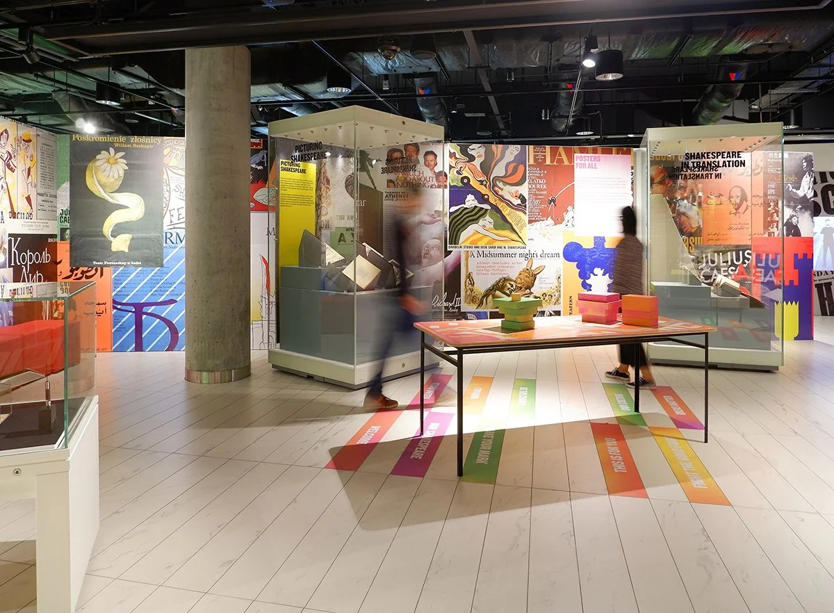 Visitors in a colourful gallery. In the background, a long wall is covered with posters for Shakespeare plays in different languages. Freestanding showcases display open books. A table in the centre is stacked with piles of coloured blocks.