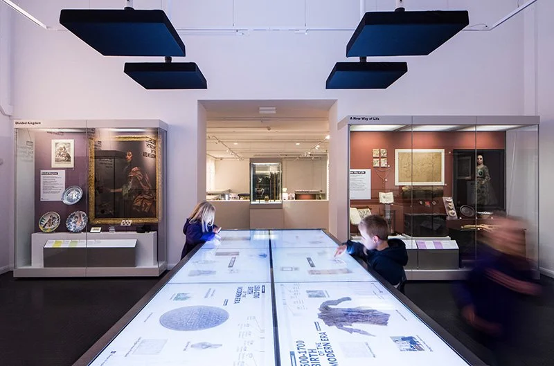 Two children stand either side of a large interactive touchscreen table exploring its content. Large showcases on both sides of the room display ceramics, artefacts and paintings. In the background, more displays are visible in a second gallery.