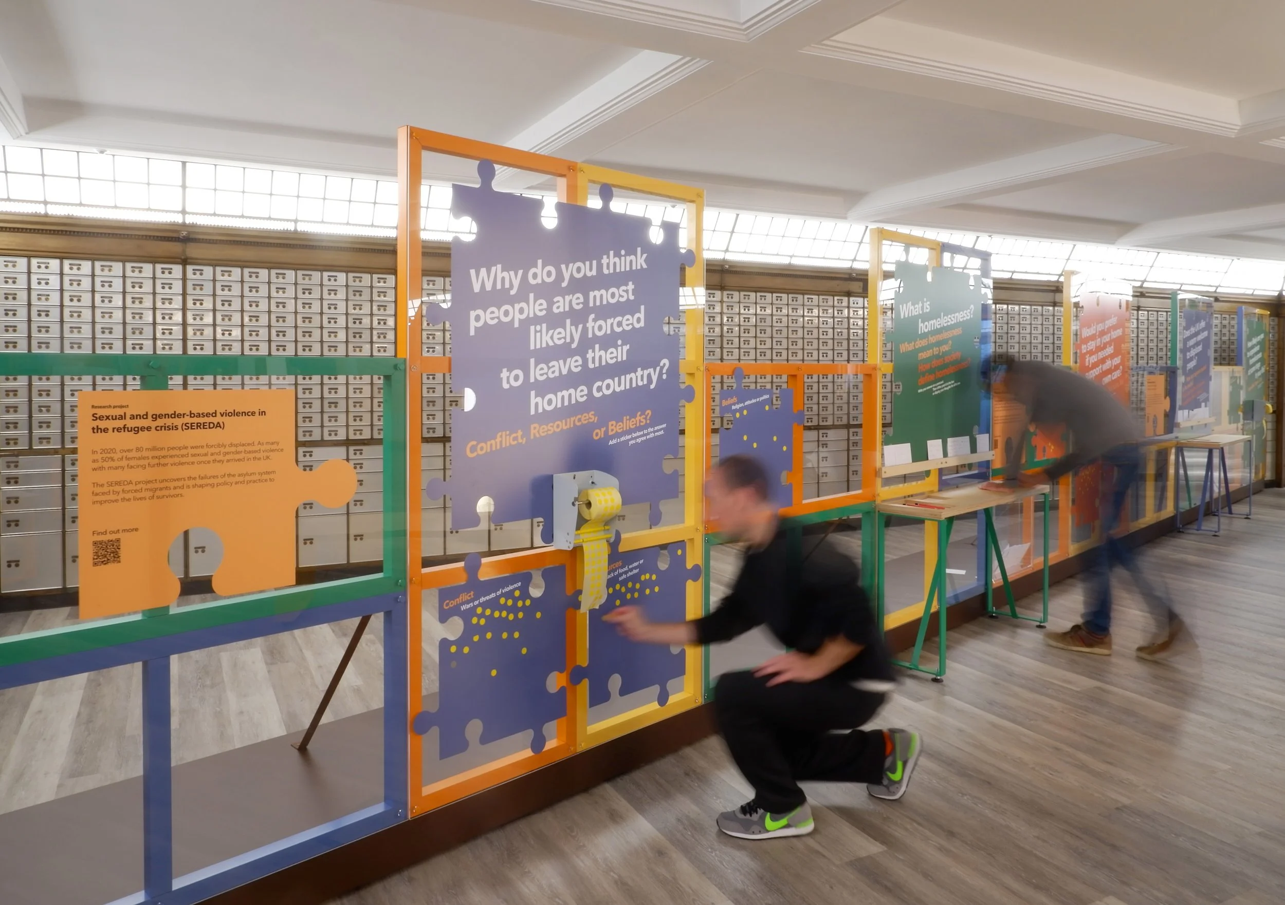 Visitors interacting with a colourful exhibition structure, with text panels in the shape of jigsaw pieces. One man kneels and places a sticker beneach a panel that asks ""Why do you think people are most likely force to leave their home country?"