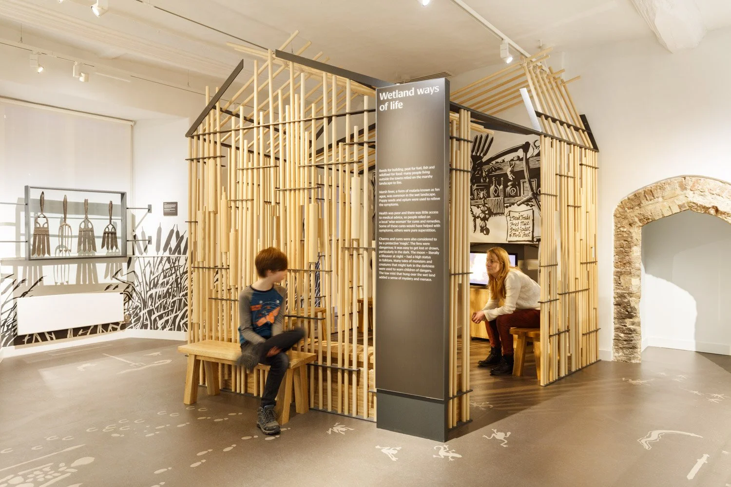 A woman sits inside a wooden structure in a gallery. Its walls are made from timber dowels, and a woodcut illustration and TV screen are visible through the walls. A title outside reads “Wetland ways of life”