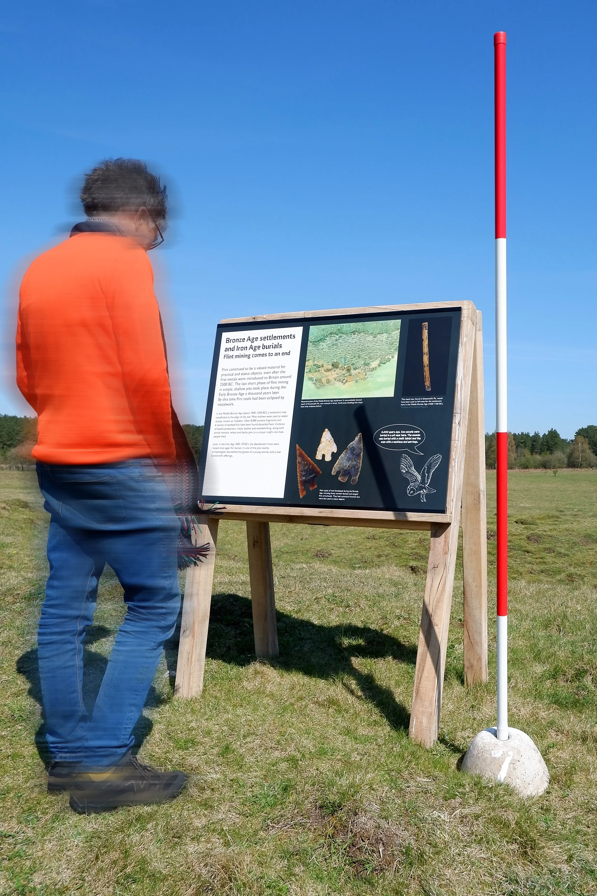 A man wearign an orange sweater walking in a field with an A-board way-finding and interpretation sign featuring an eagle illustration, Neolithic tools, and way-finding information.