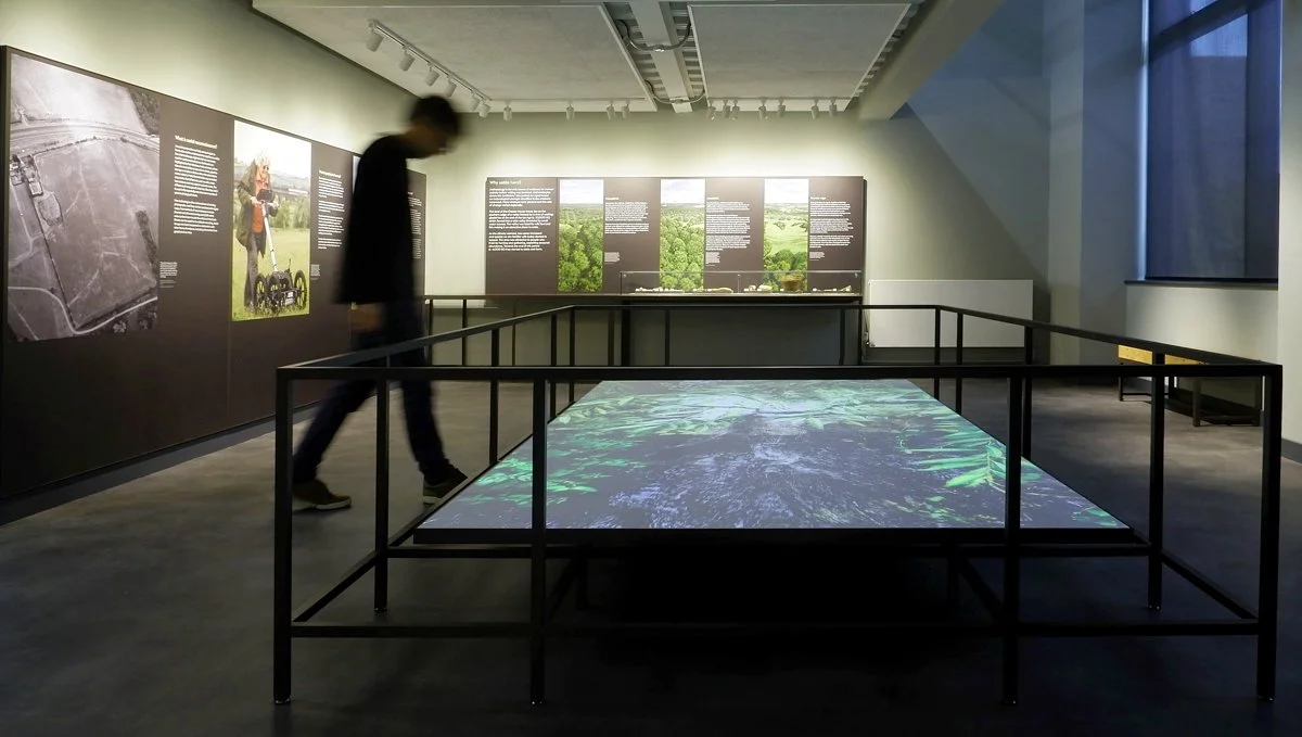 In a gallery a man looks down at a large projection of greenery at floor level, surrounded by a black railing. Interpretation panels line the left and far walls, with large images of landscape and white explanatory text.