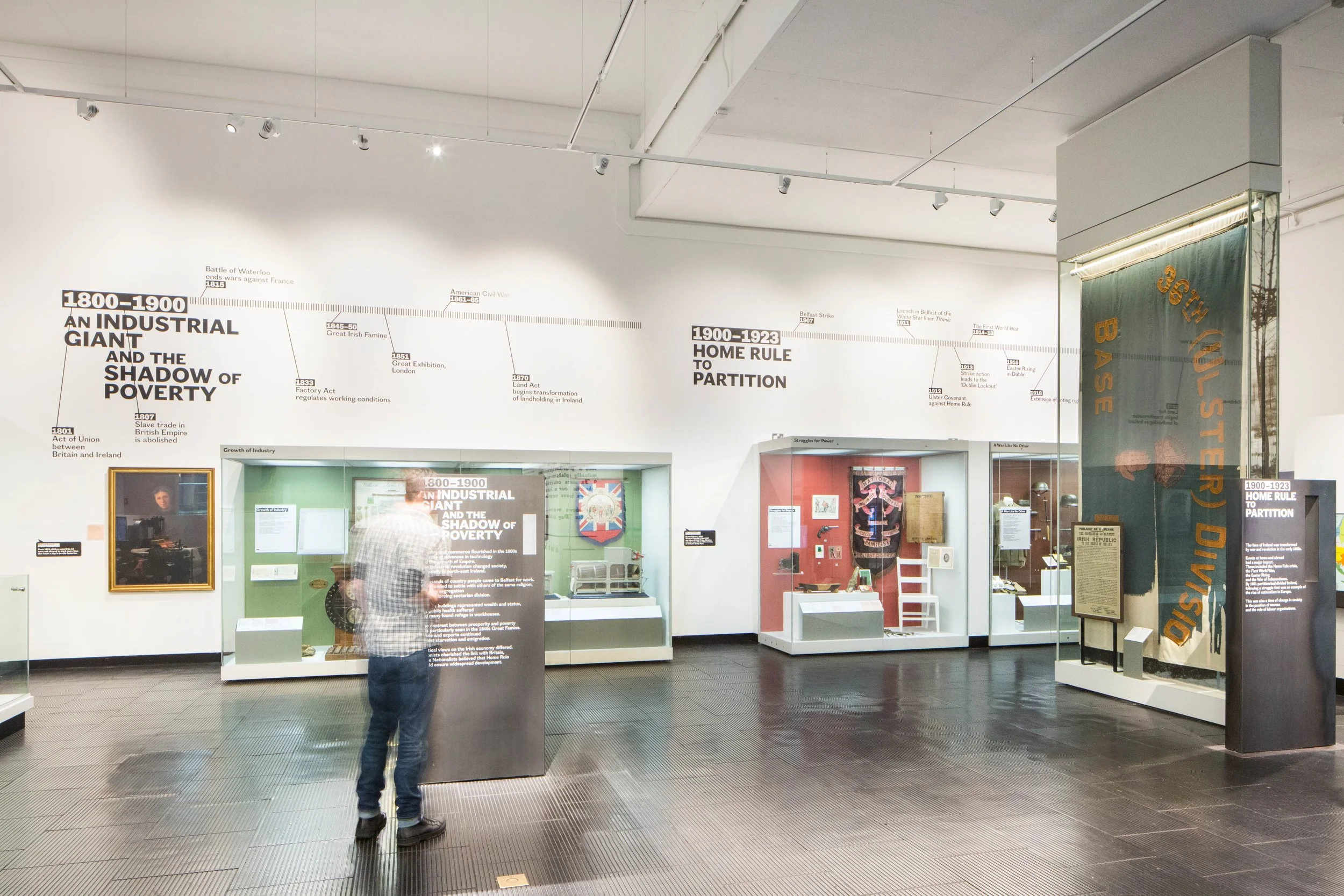 A visitor reads a text panel in a gallery. Showcases displaying artefacts line a white wall, and above them a large timeline provides key dates between 1800-1923. To the right, a large green flag is displayed in a freestanding case.