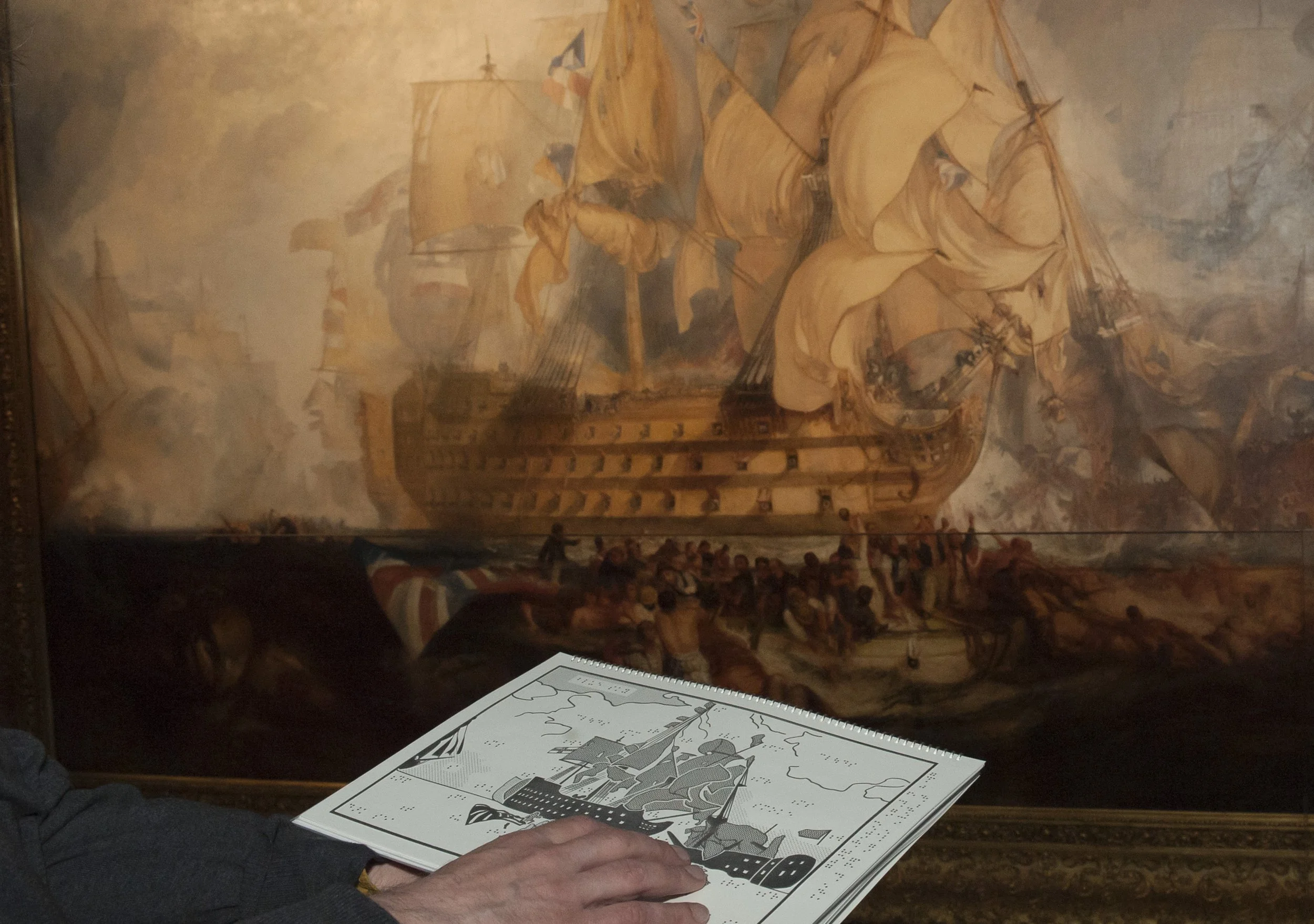 A person touches a tactile image of The Battle of Trafalgar in a printed booklet, in front of the painting itself. The booklet is accompanied by braille and a large print guide.