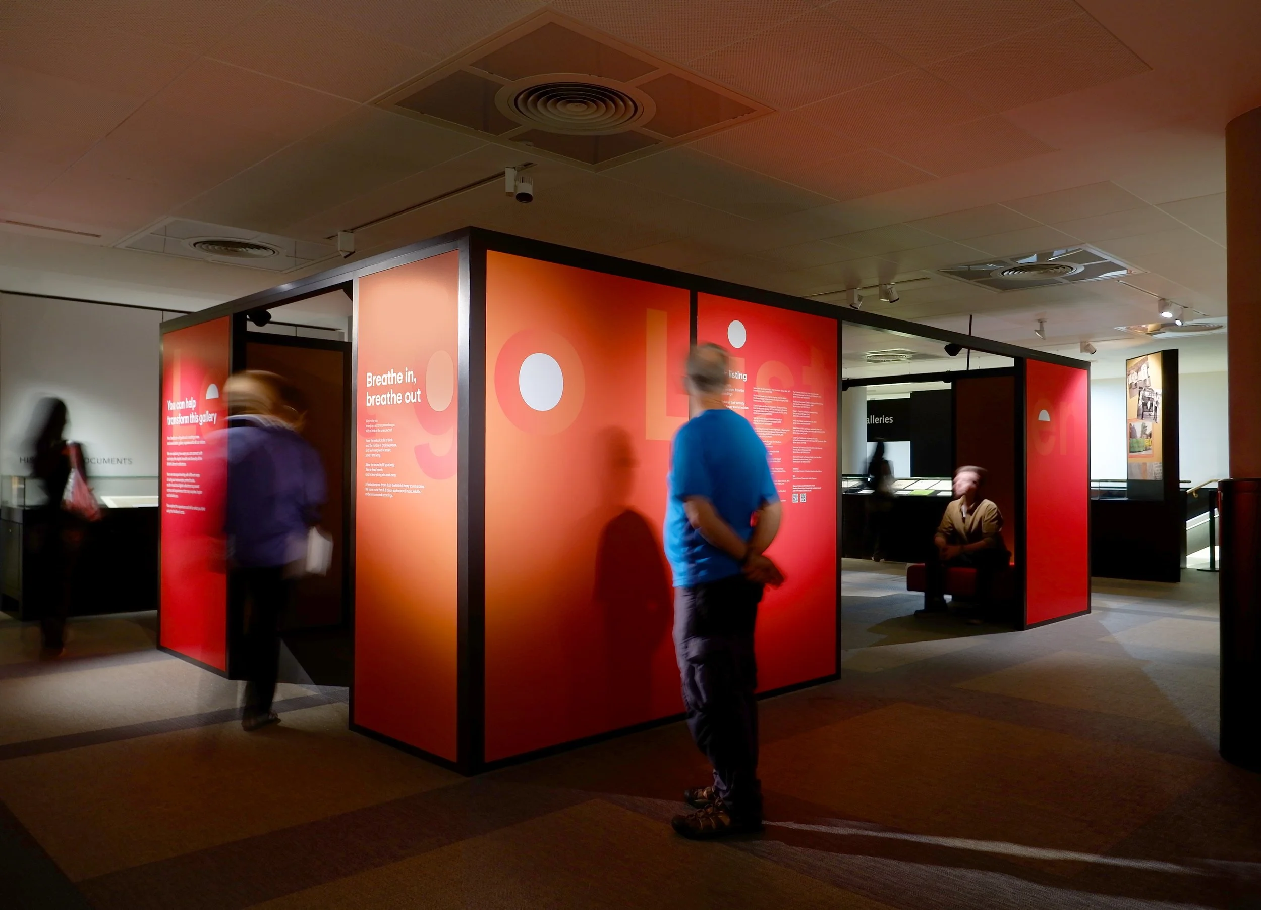 A rectangular structure stands in the middle of a gallery at the British Library. The graphic surfaces of the structure are red and carry textual information. A man enters from one of the entrances, while a second man sits on a red bench.