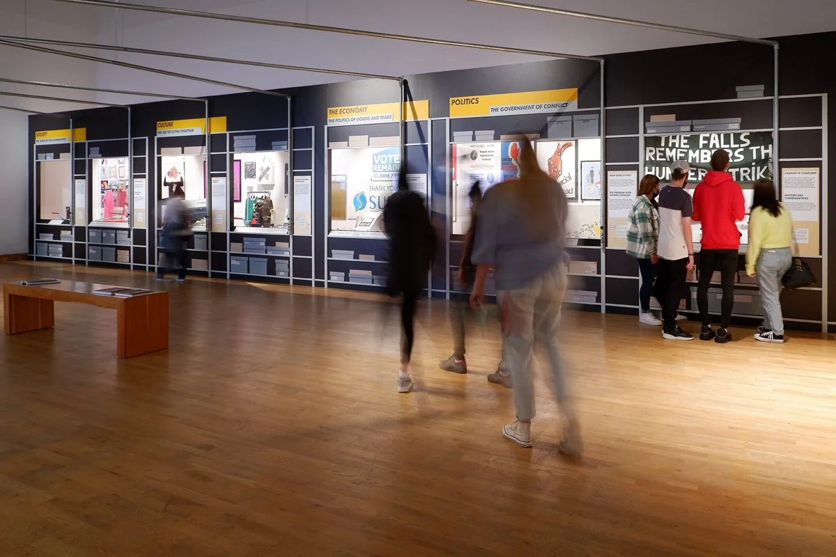 Groups of visitors look at displays in a gallery. Each display section has a yellow title, a showcase display and graphics of storage boxes beneath it. Metal tubes run from the display across the ceiling.