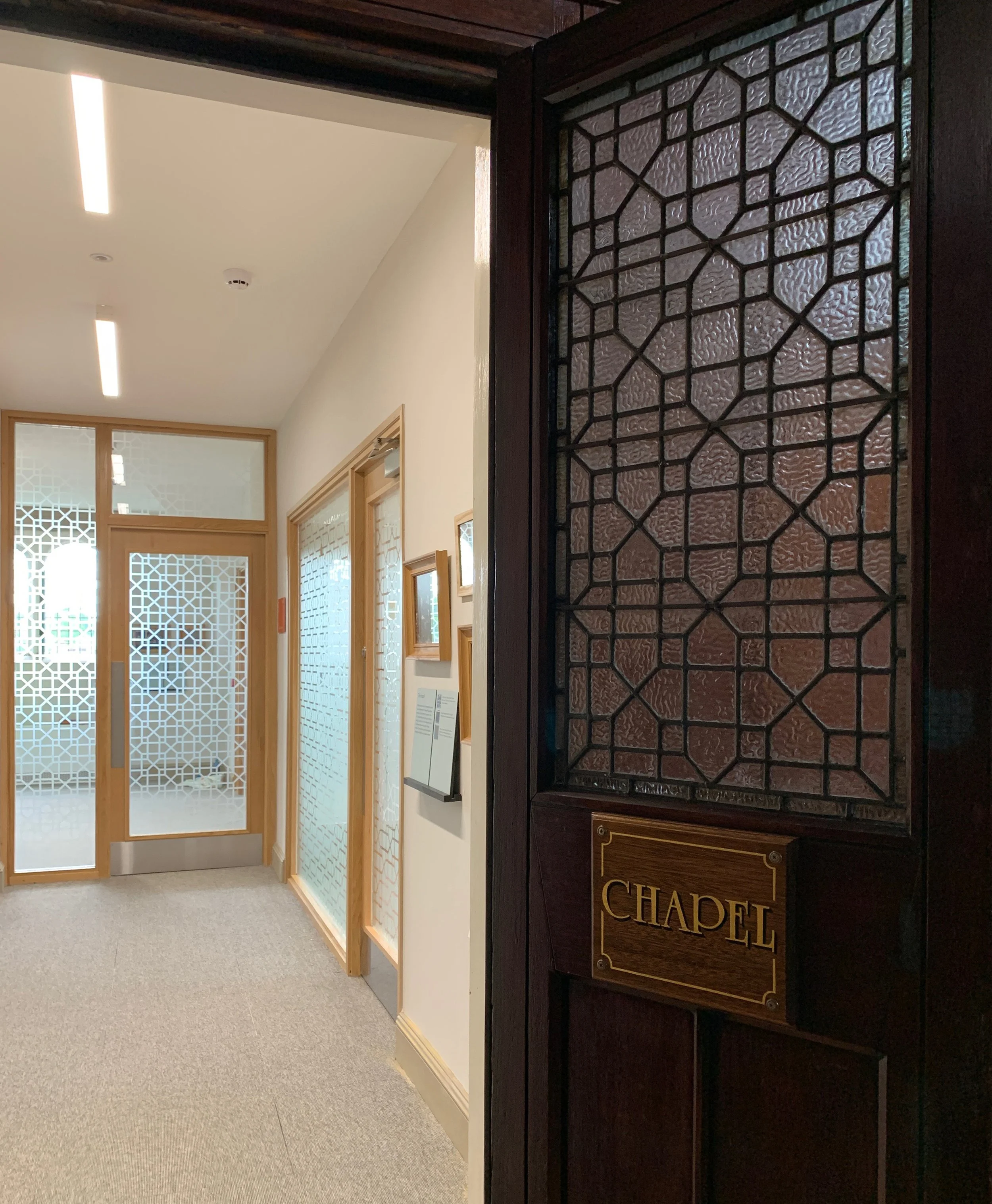 A dark wooden door with a frosted window panel and a wooden sign reading “Chapel” stands open, leading into a modern hallway with glassed doors and walls leading to other rooms.