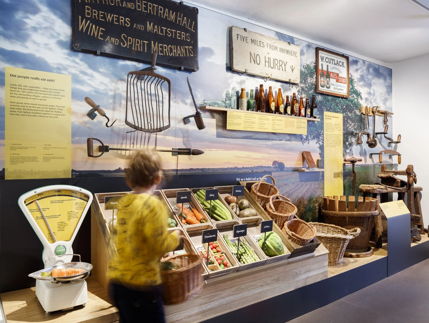 A display of market produce in a gallery against a large image of a field. A young child holding a basket walks past a weighing machine and wooden vegetables in crates.