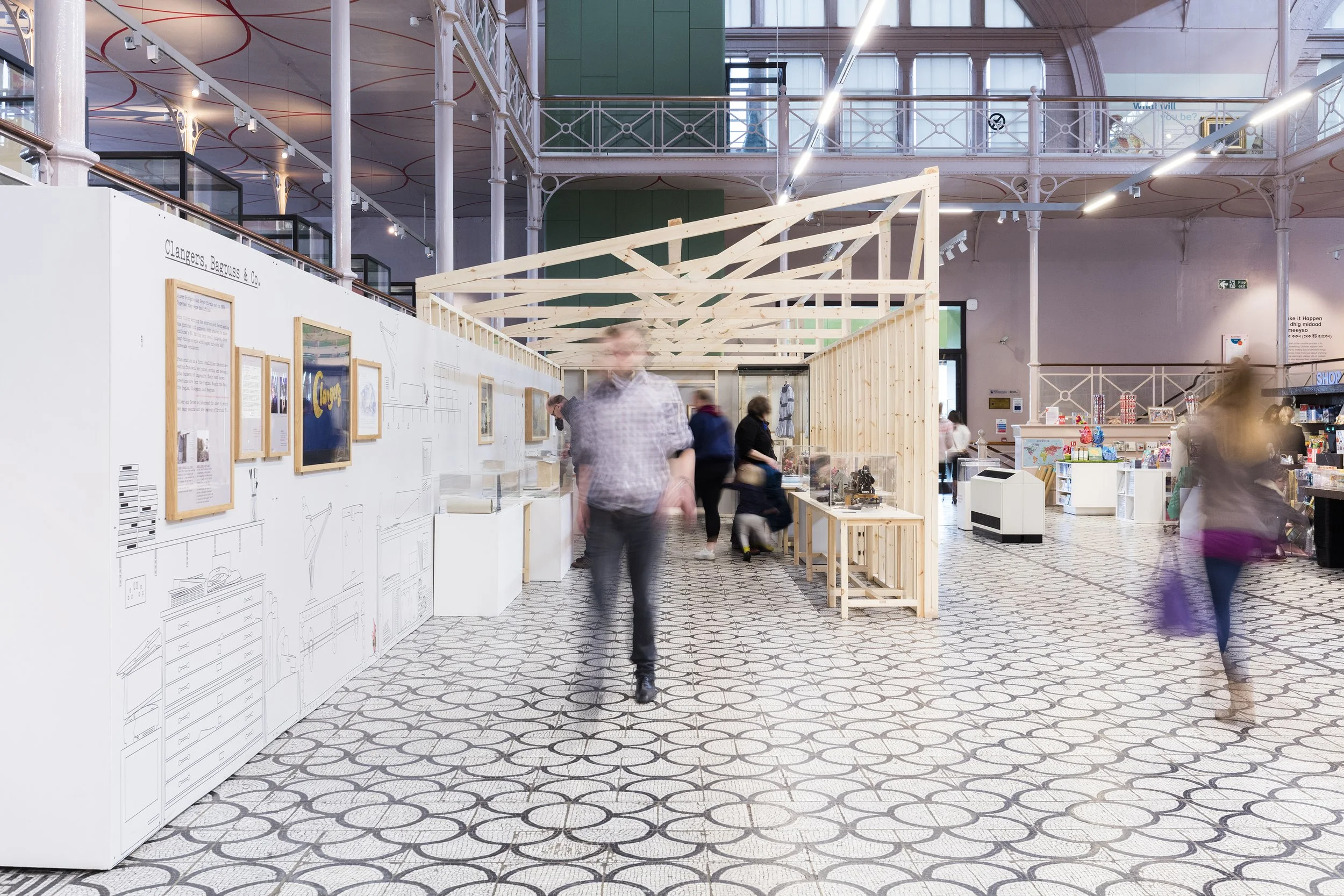 The entrance to a minimal timber structure with visitors looking at objects inside. On the left of the space, a white wall displays the title "Clangers, Bagpuss & Co" with line drawings of a creative workspace and framed photographs.